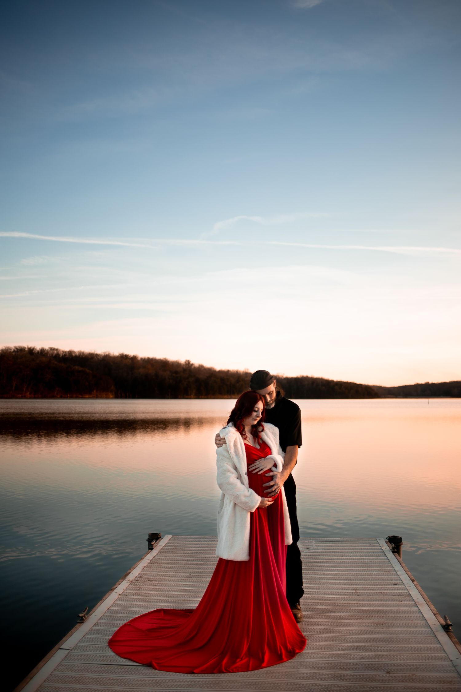 Couple shares intimate moment on wooden dock by peaceful lake at sunset, bride wearing flowing red dress.