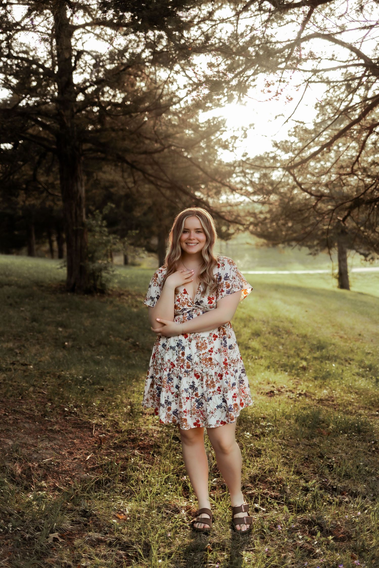 Young woman in a floral dress poses in a sunlit park setting.