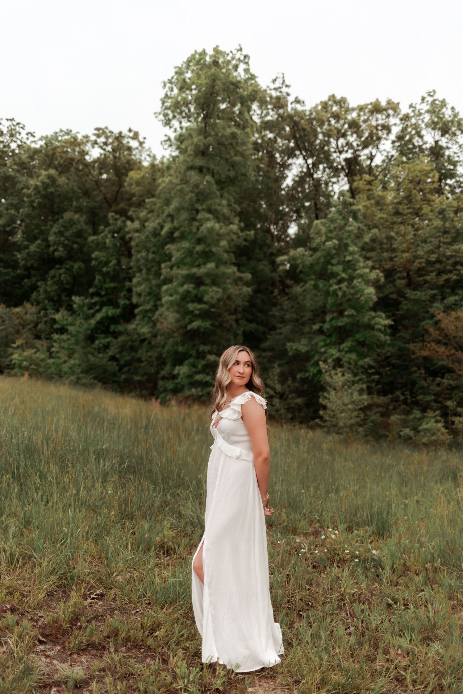 Ethereal outdoor portrait in white dress against green meadow and forest backdrop.