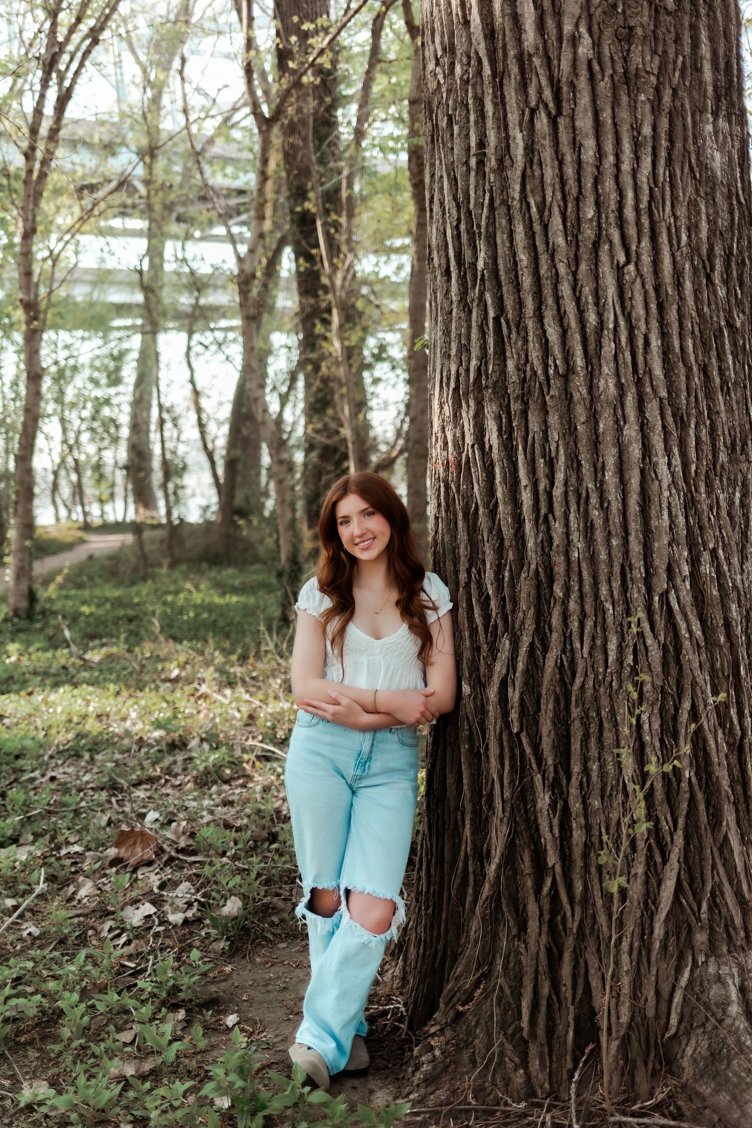 Person leaning against large tree trunk wearing ripped jeans and white top near a lake.