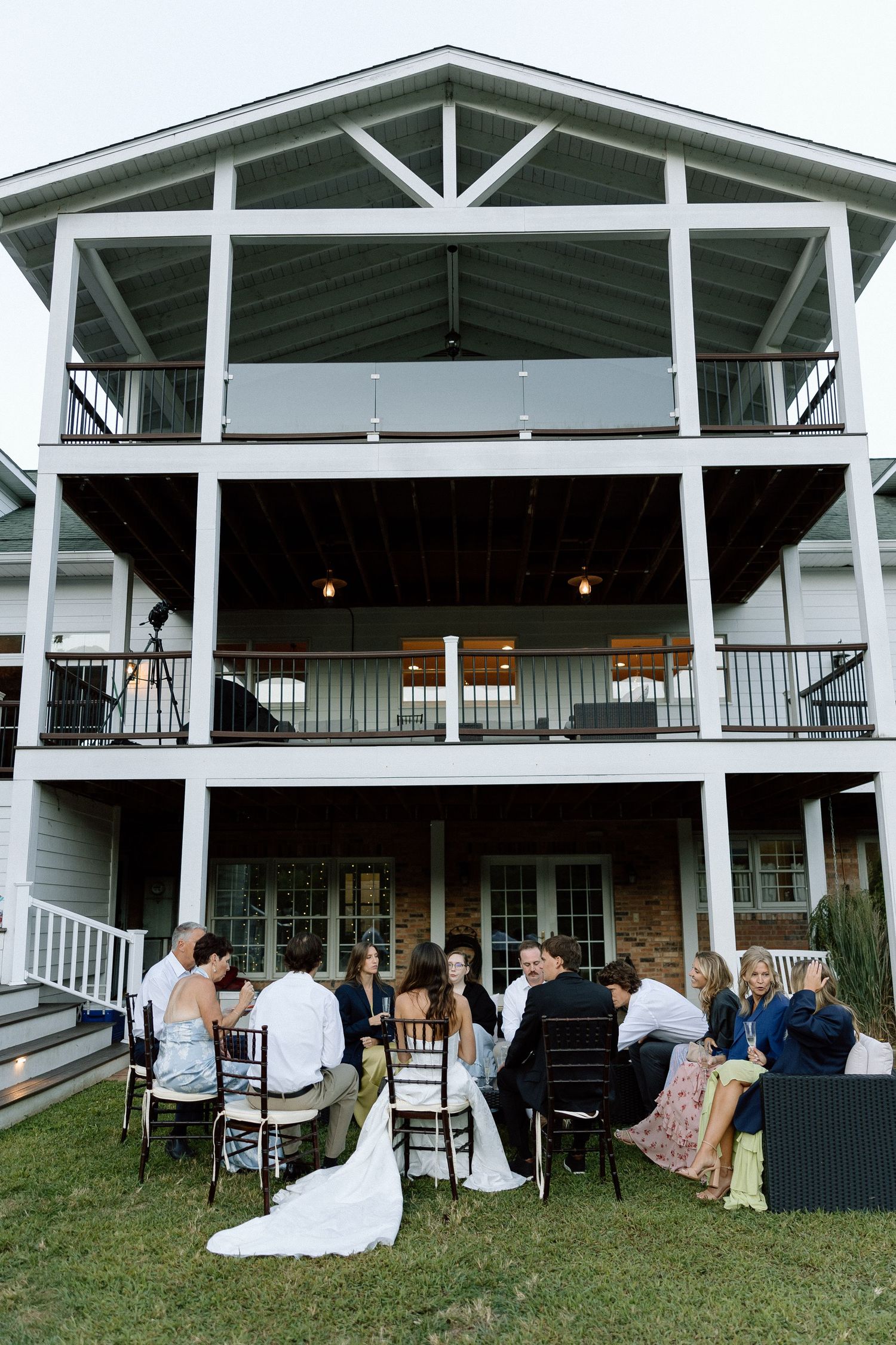 Guests gather for an outdoor reception on the lawn of a three-story white house with wrap-around porches and balconies.