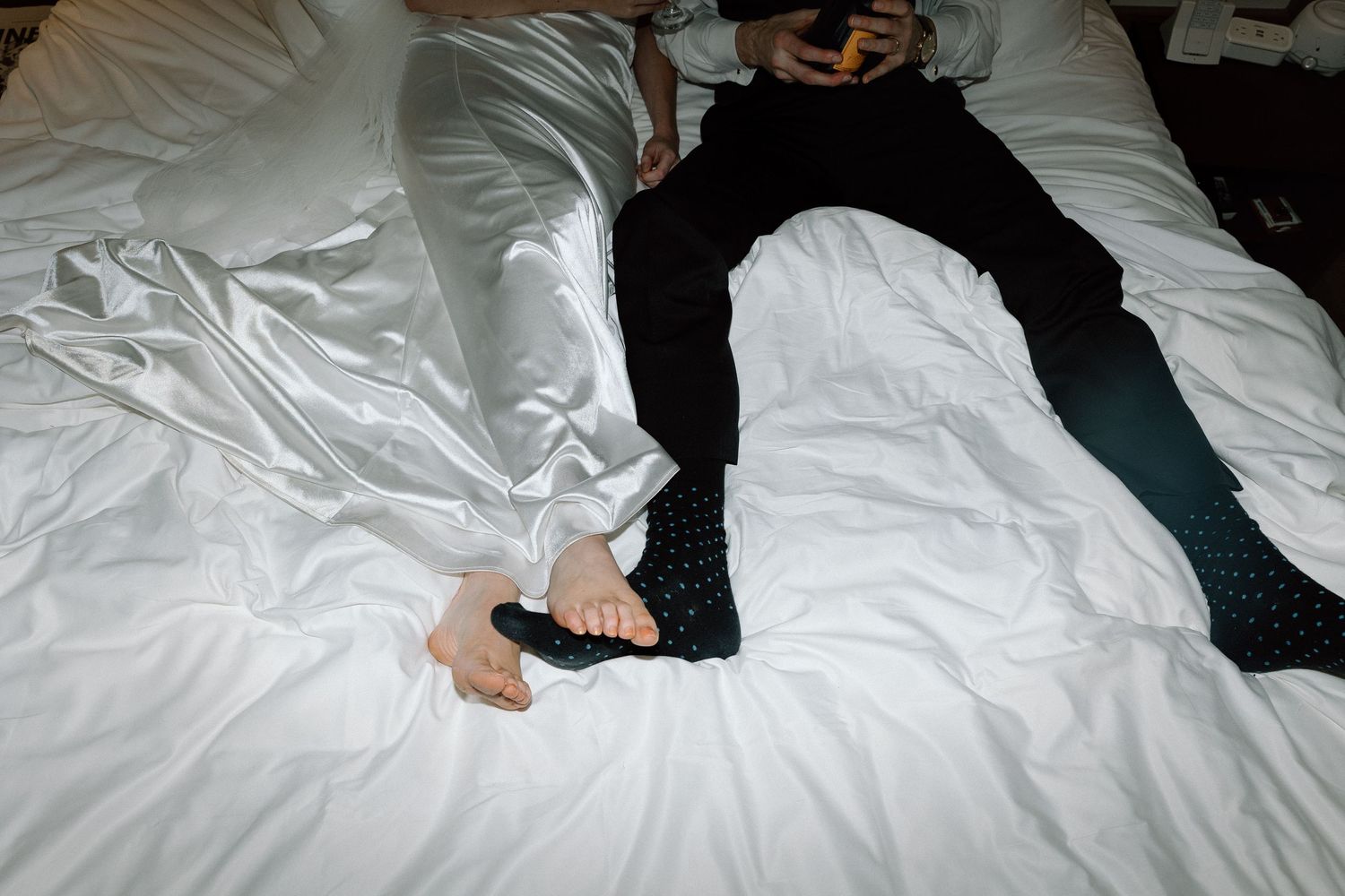 Someone in dark clothing lies on white bedding while wearing casual clothes and shoes.