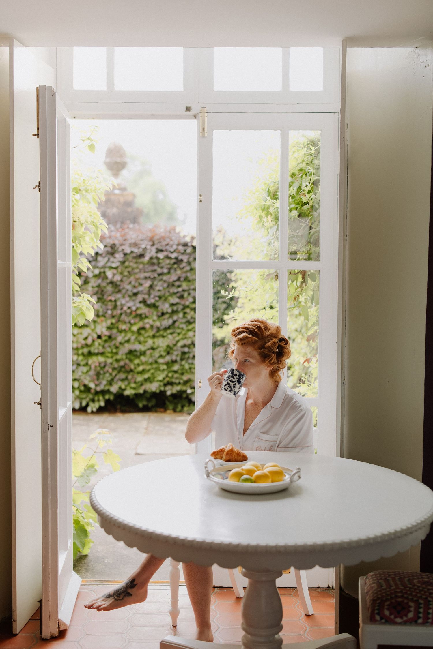 Someone in a white robe enjoys breakfast at a round white table by French doors overlooking a sunny garden patio.