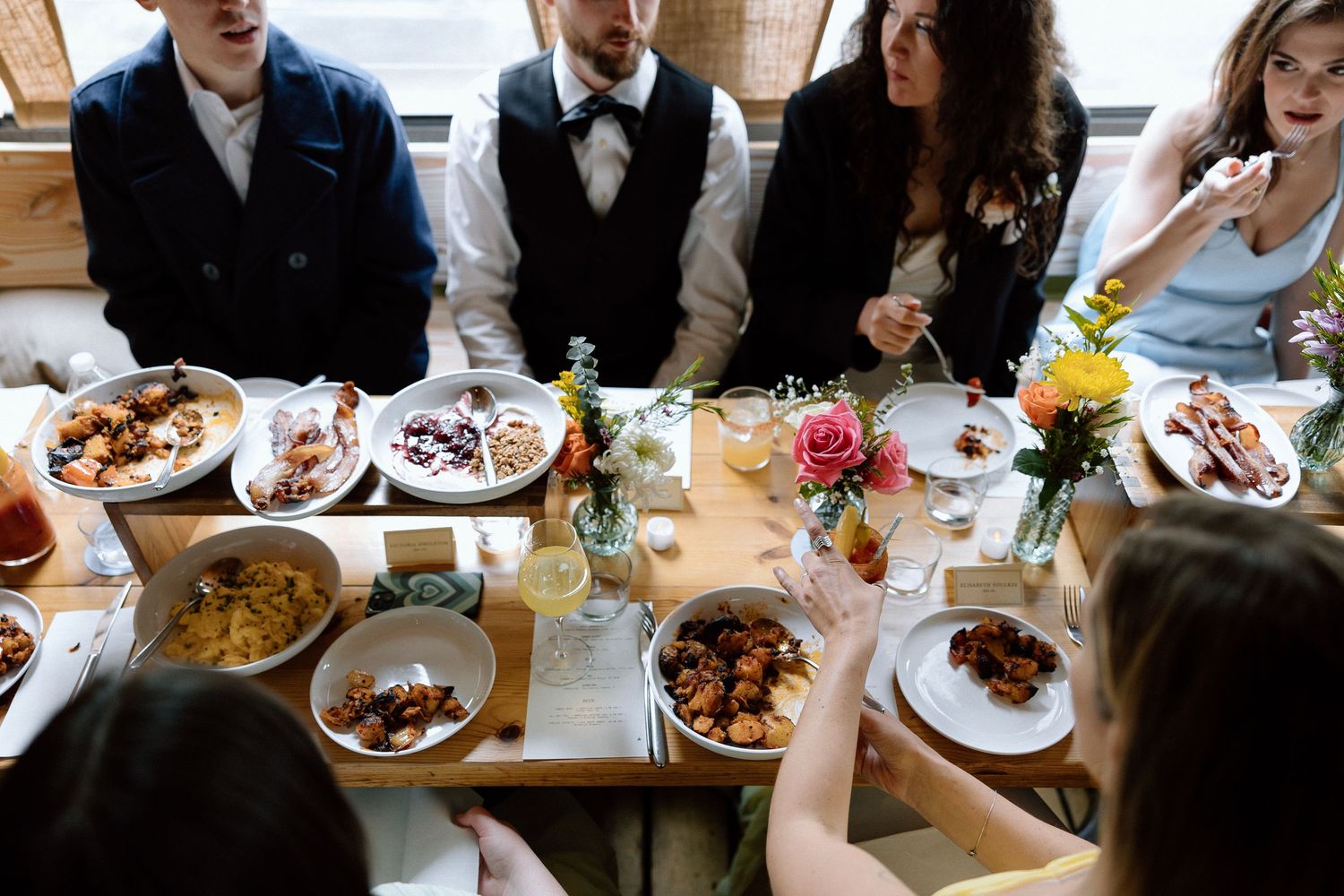 A group of people gather around a wooden dining table filled with plates of food and floral centerpieces.