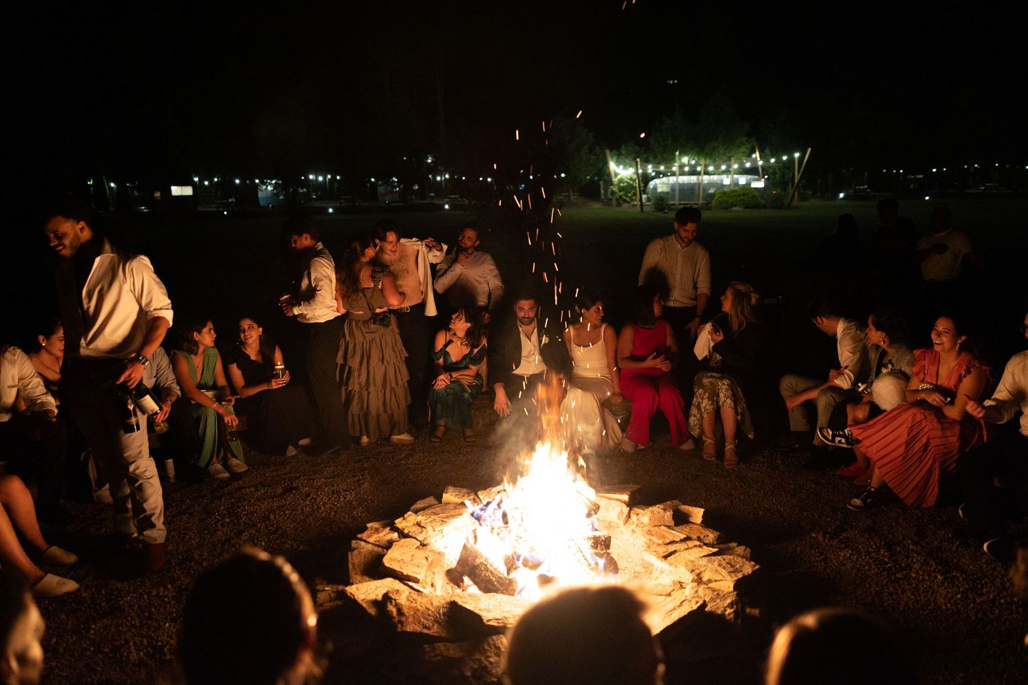 A group of people gathered around a large bonfire at night with city lights visible in the background.