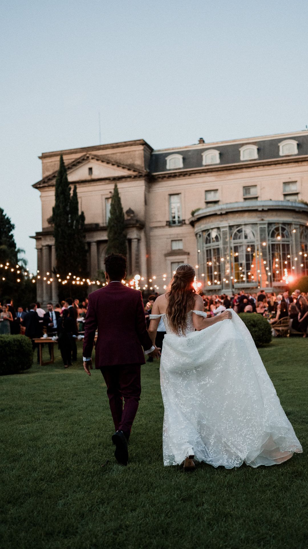 Couple walking on lawn towards elegant venue with string lights at dusk during outdoor wedding reception.
