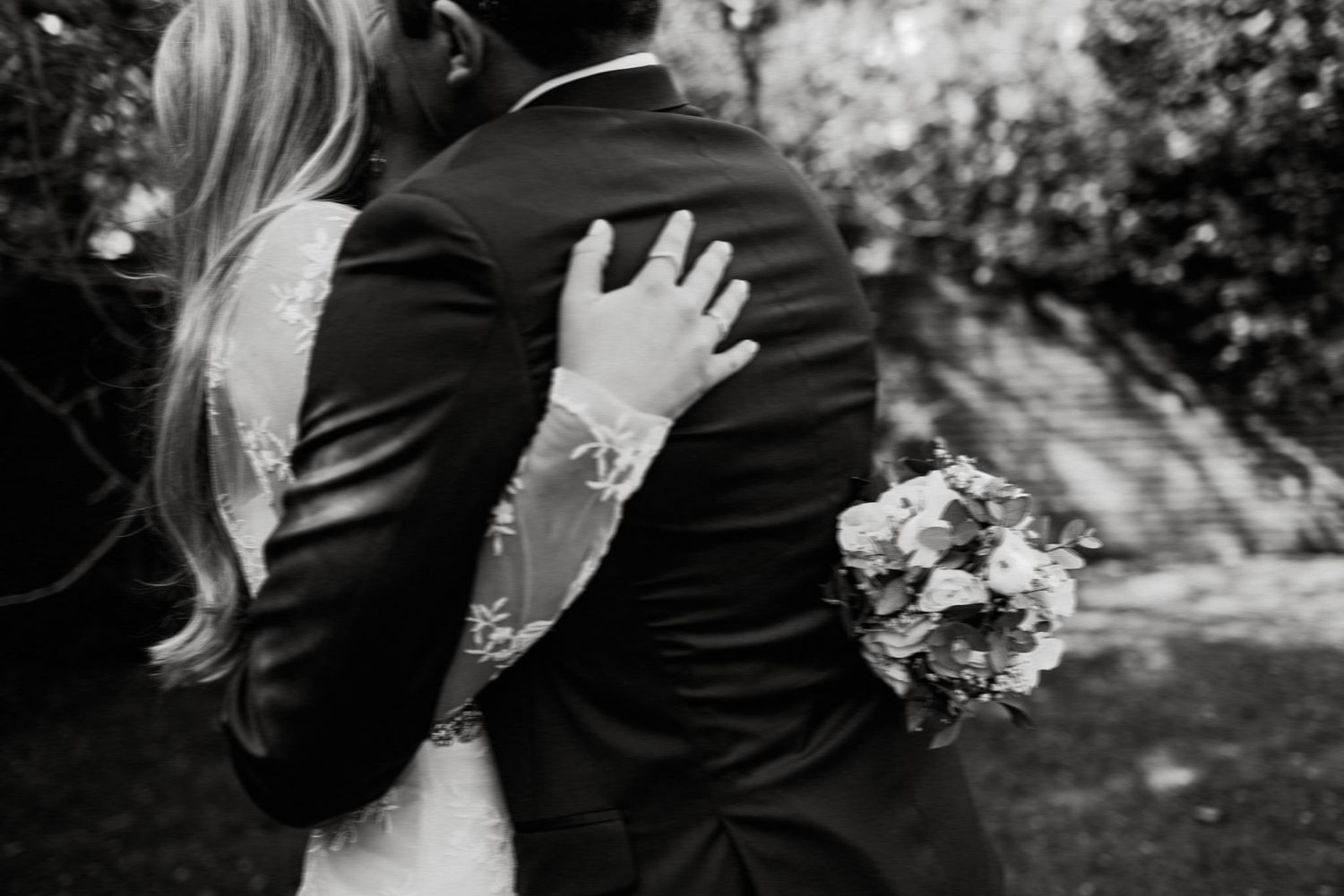 Black and white photo showing a close-up embrace between newlyweds, with the bride holding a floral bouquet.