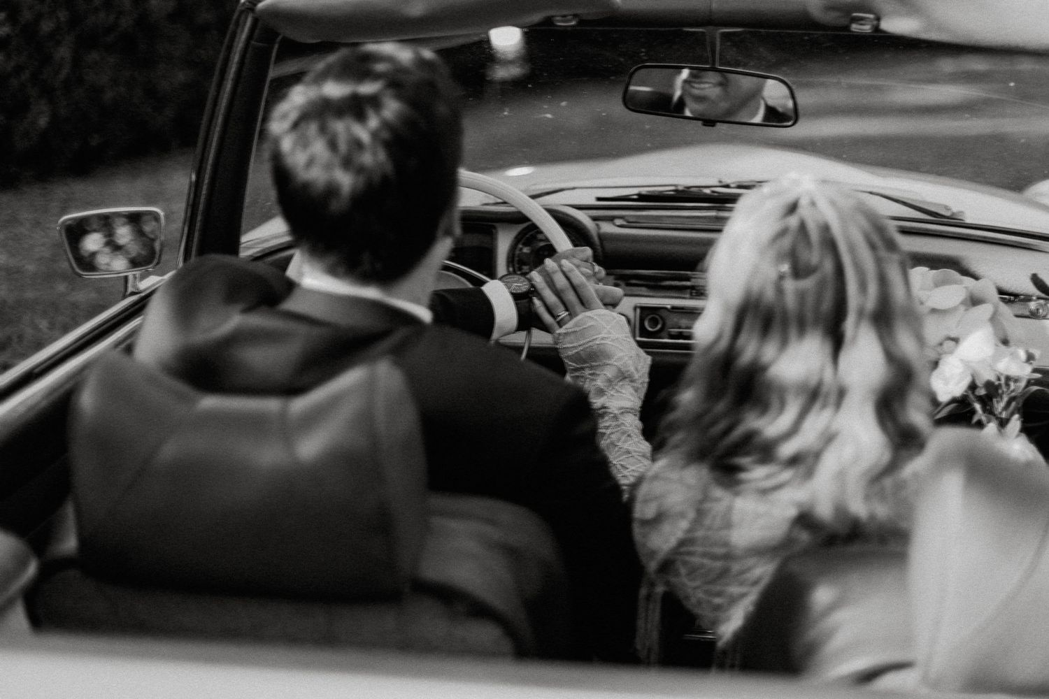 A black and white view from behind of two people riding in a classic convertible car on a sunny day.
