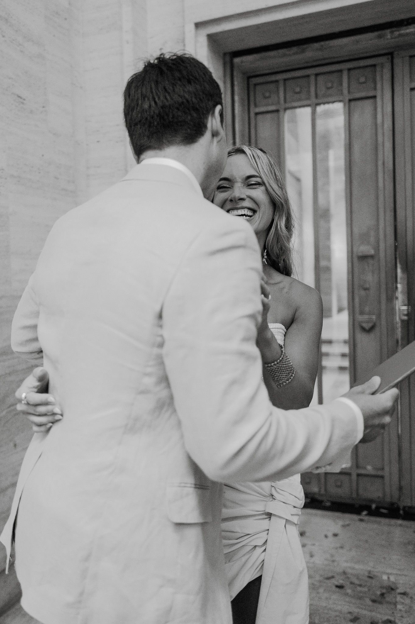 A couple embraces in a candid black and white wedding moment in front of a doorway, showing joyful emotion and connection.