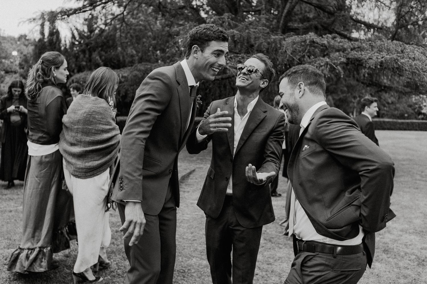 Three men in suits share a candid laughing moment at an outdoor gathering.