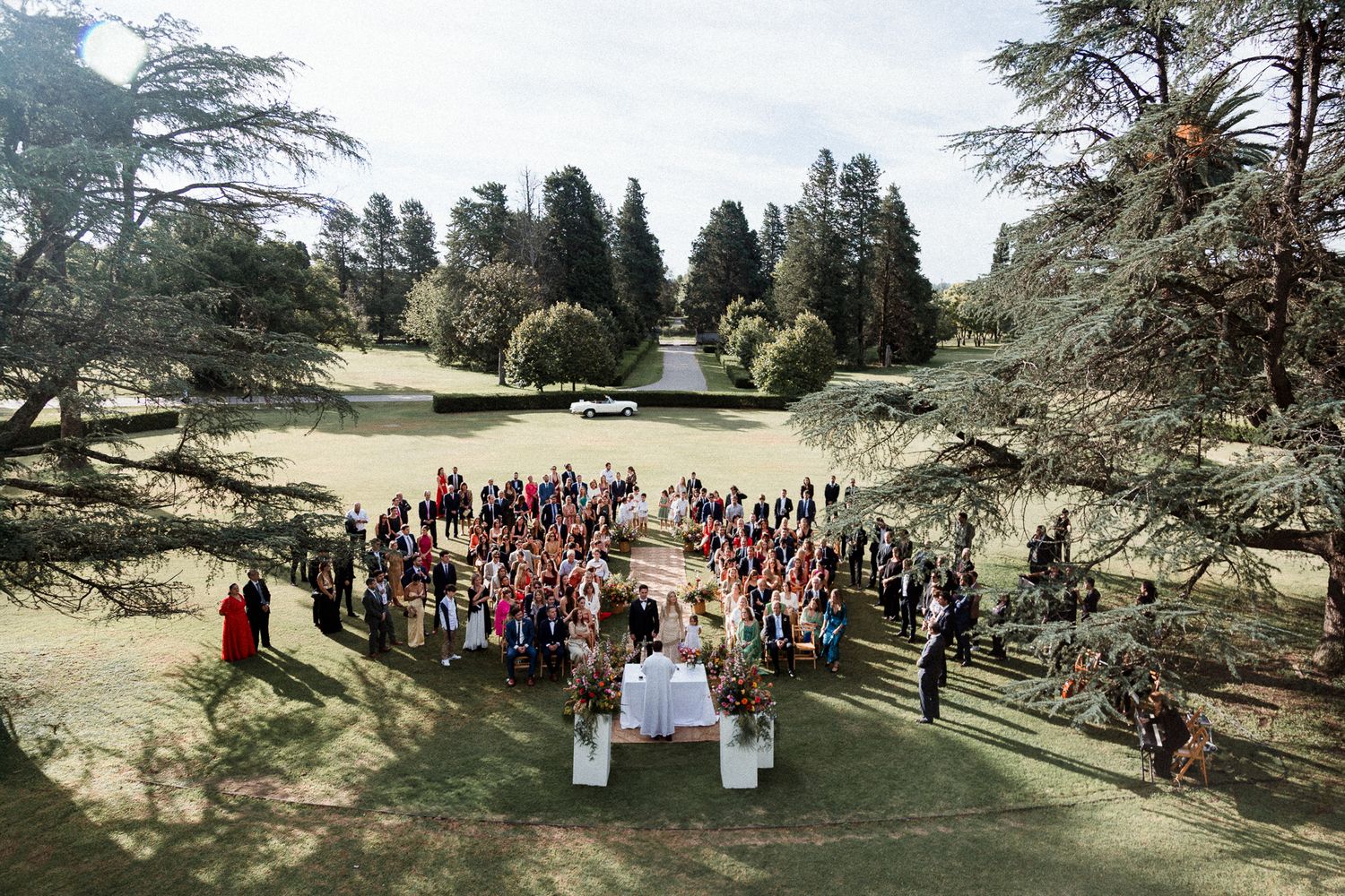 Aerial view of an outdoor wedding ceremony surrounded by tall trees in a lush garden setting during daytime.