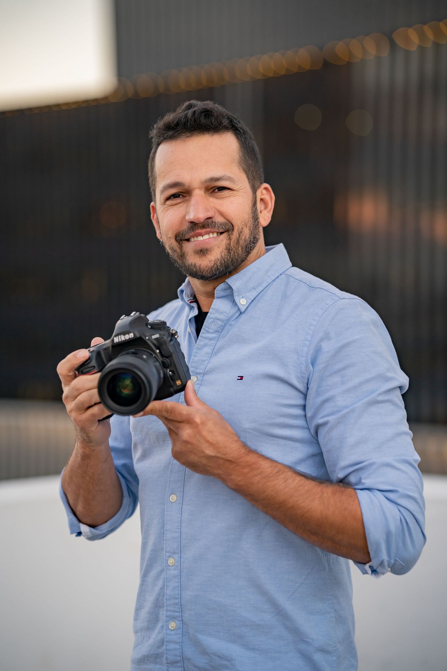 Photographer smiling at camera while holding DSLR in casual business attire.