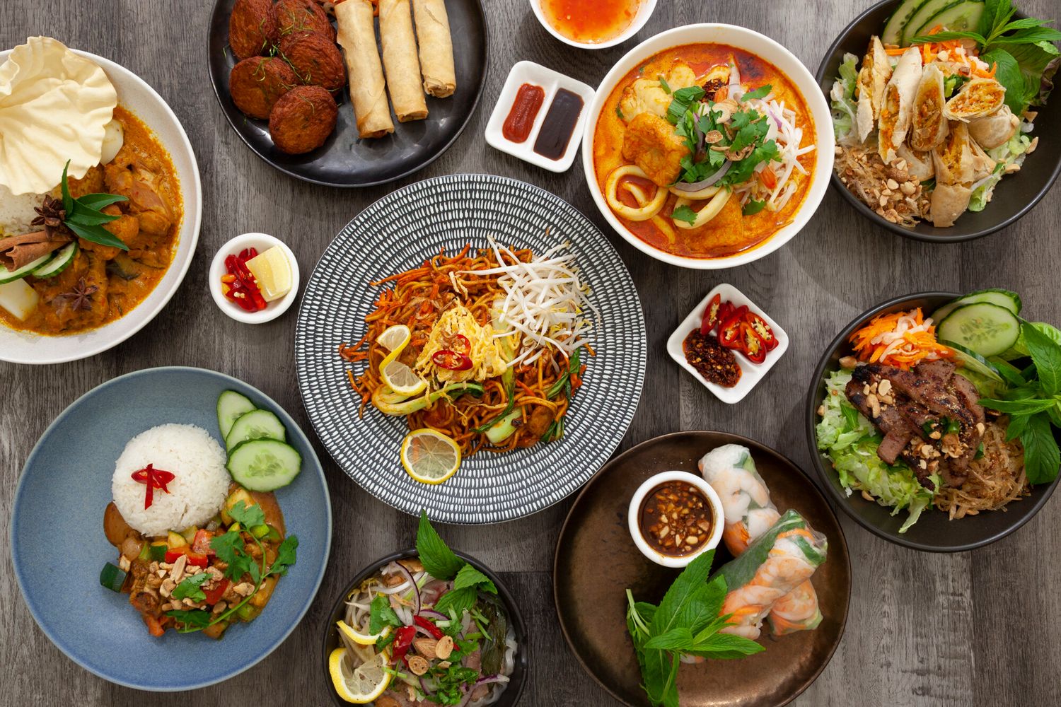 Overhead view of various Asian dishes including soups, noodles and appetizers on dark table setting.