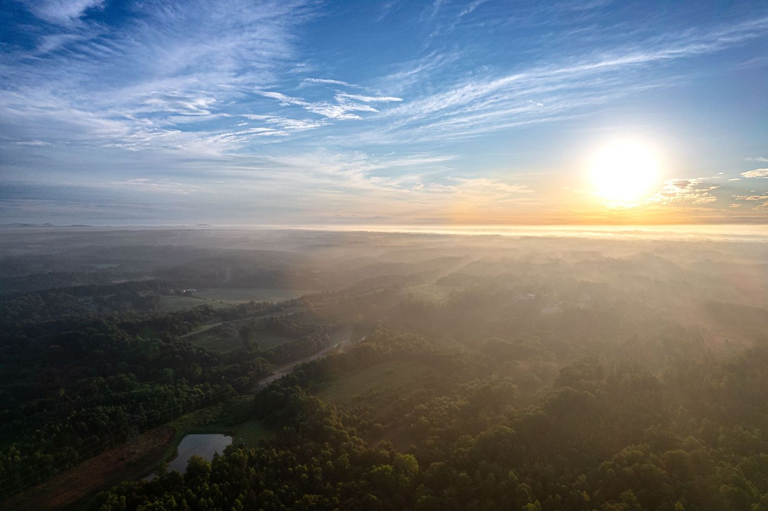 Aerial view of a misty forest landscape at sunrise with a bright sun and wispy clouds in a blue sky.