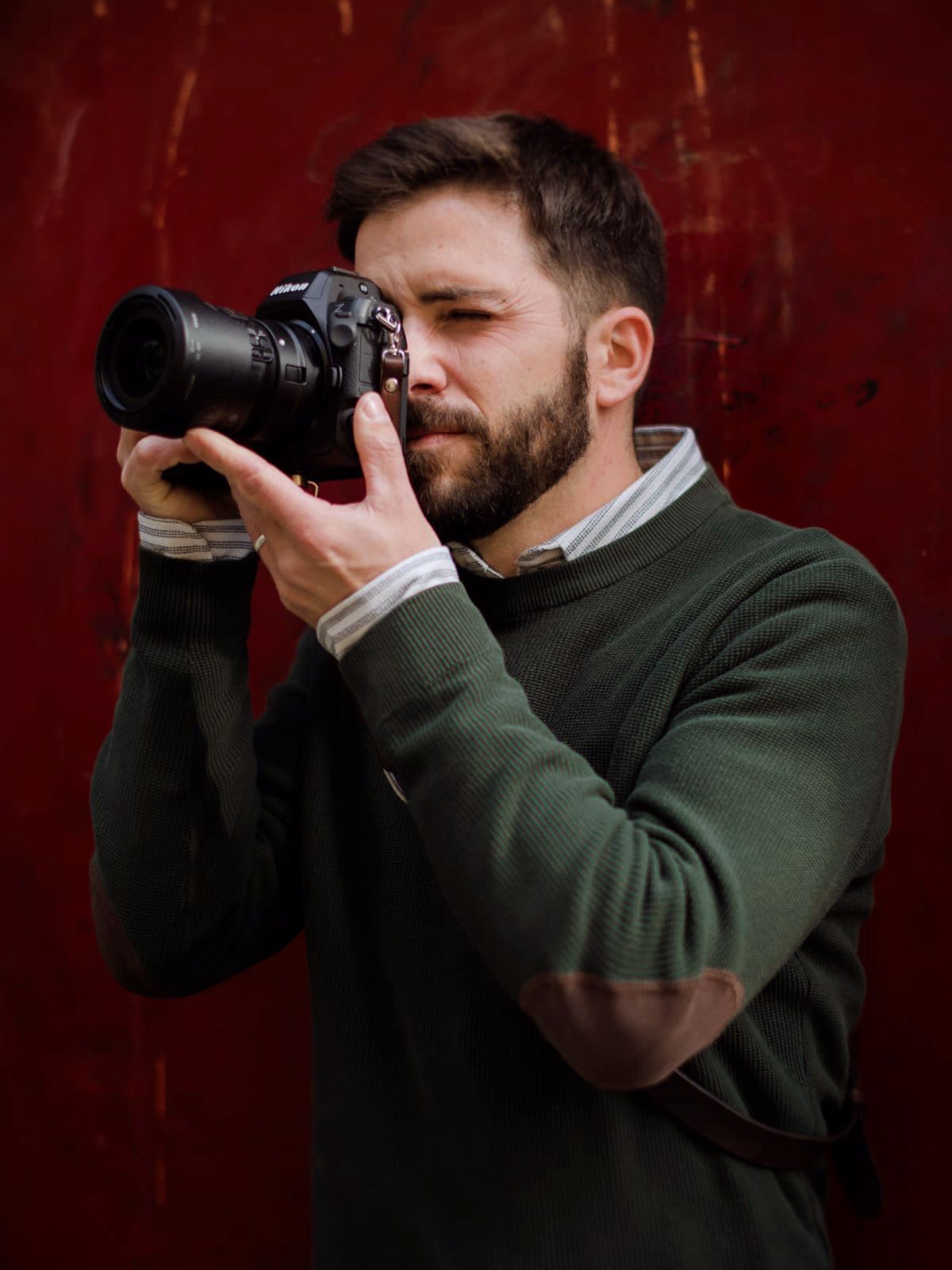 A photographer in a dark green sweater holds a professional camera with telephoto lens against a red textured background.