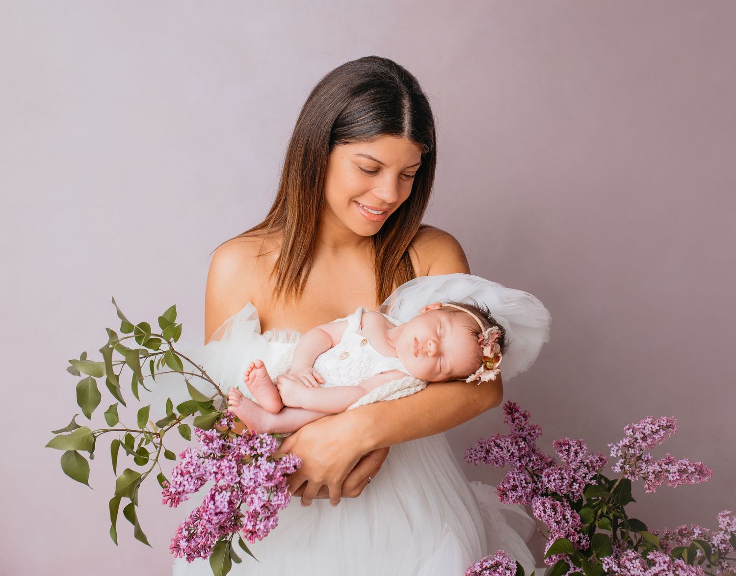 Mama in einem weißen Tüllkleid hält neugeborenes Mädchen im Arm sowie Fliederblüten ind pink