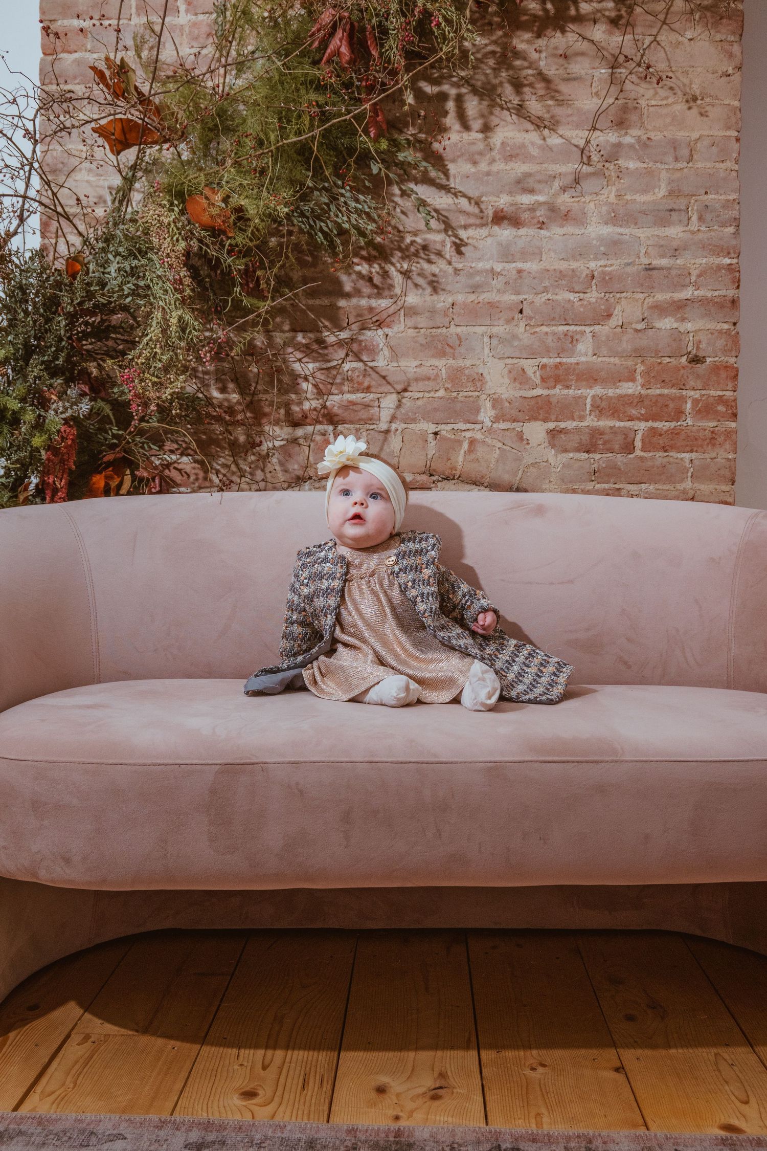 Small child in knit sweater sitting on pink curved sofa against brick wall with plants.