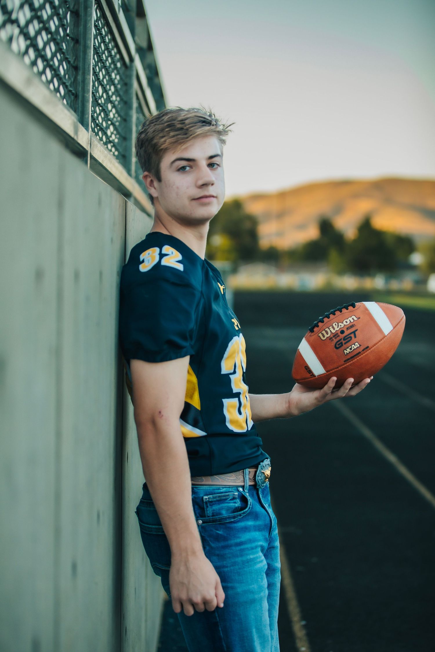 High school football player in blue jersey poses with football against metal fence.