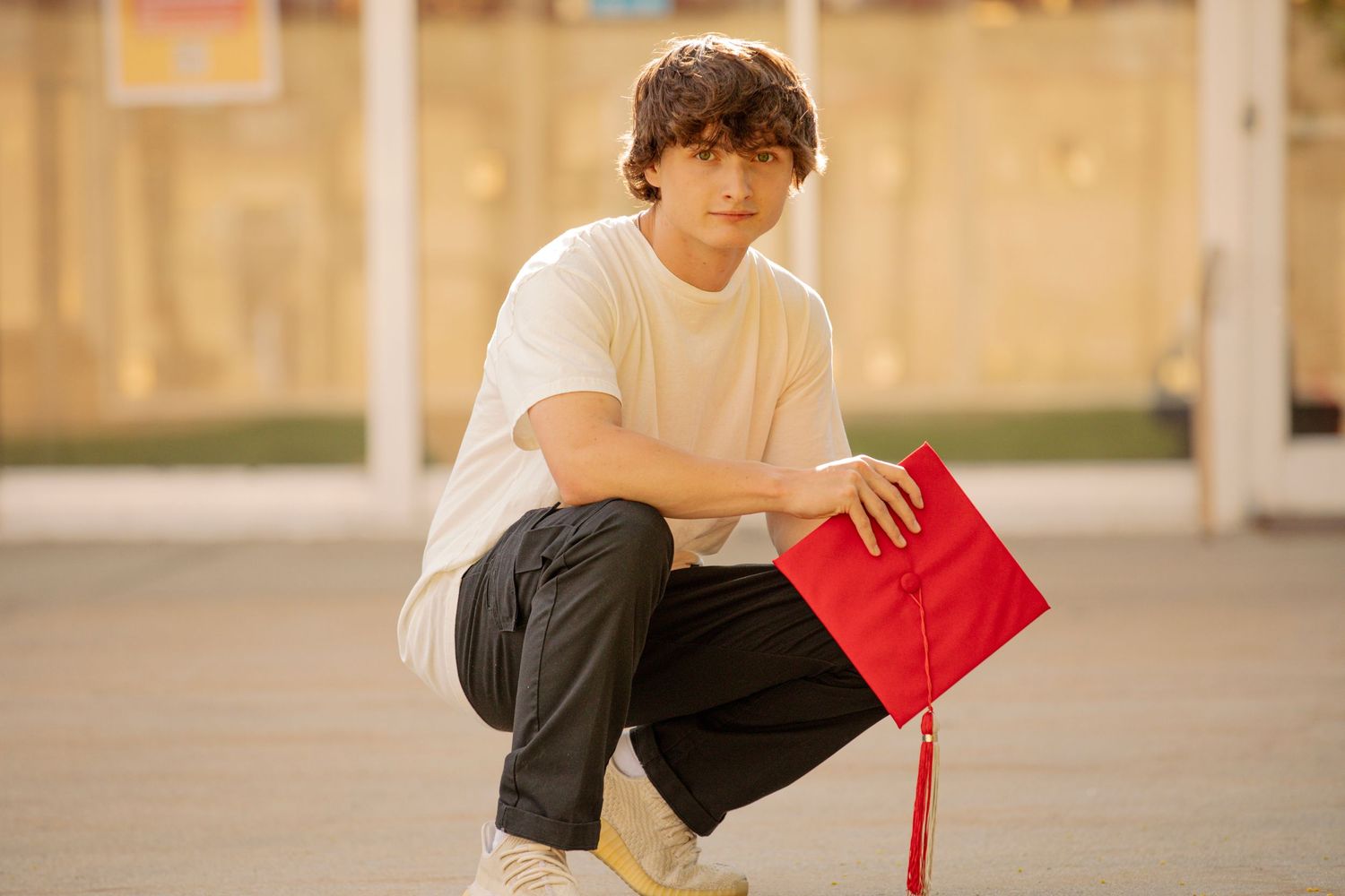 A person kneeling on concrete while holding a red graduation cap.