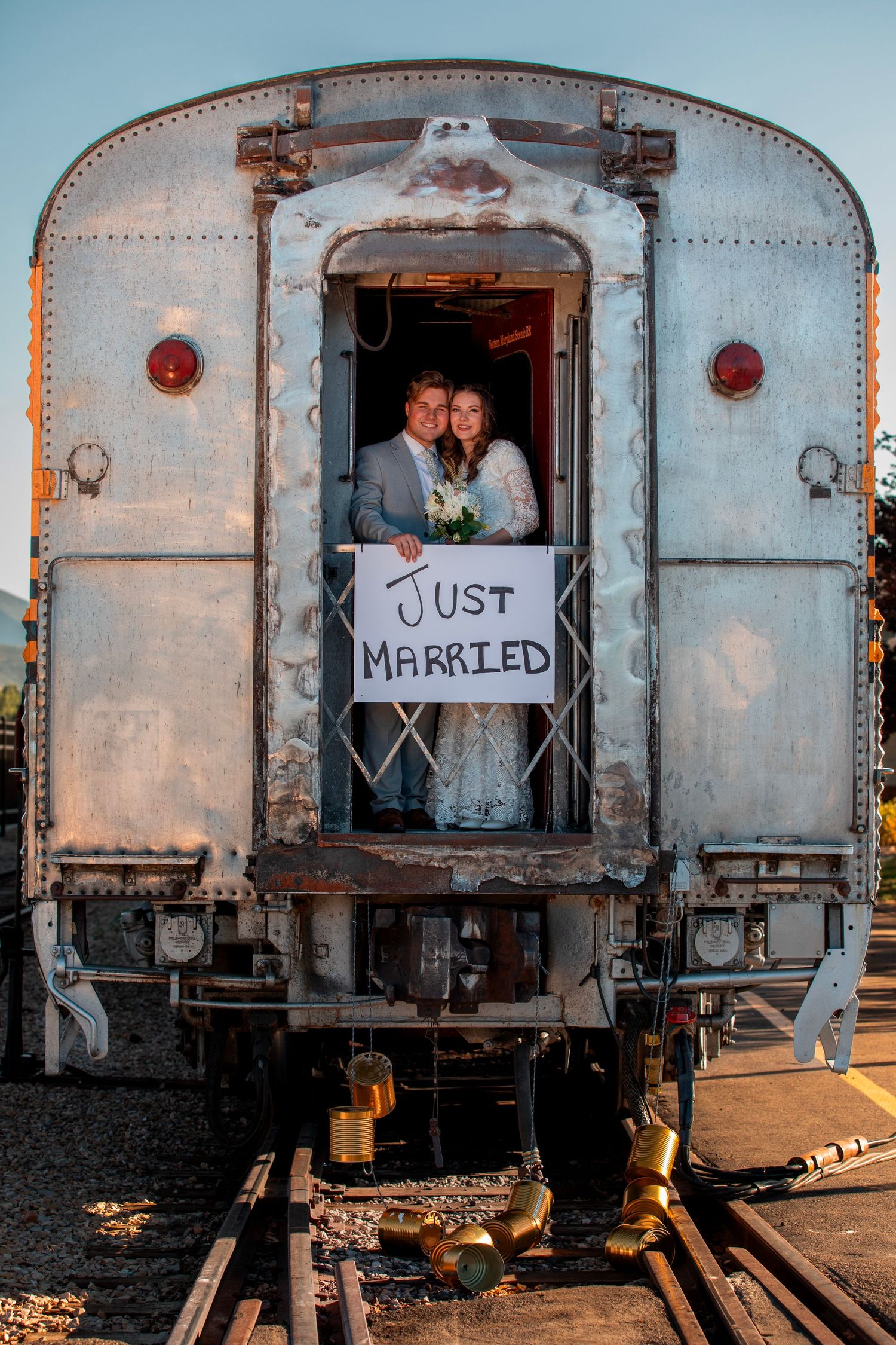 Newlyweds pose in vintage train car doorway with Just Married sign at sunset.