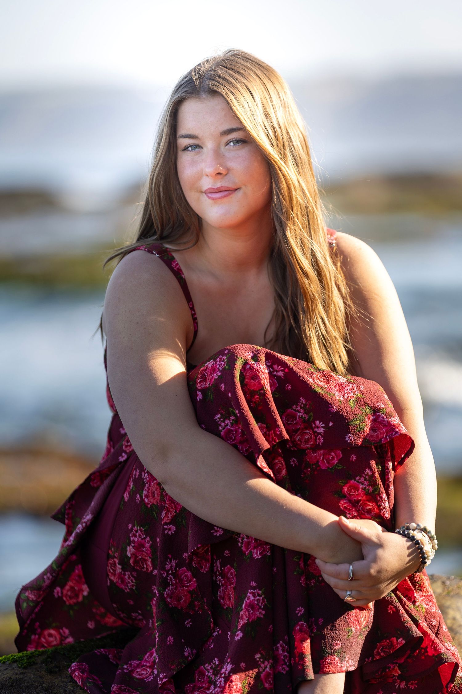 A woman in a burgundy dress poses gracefully by the water at sunset.