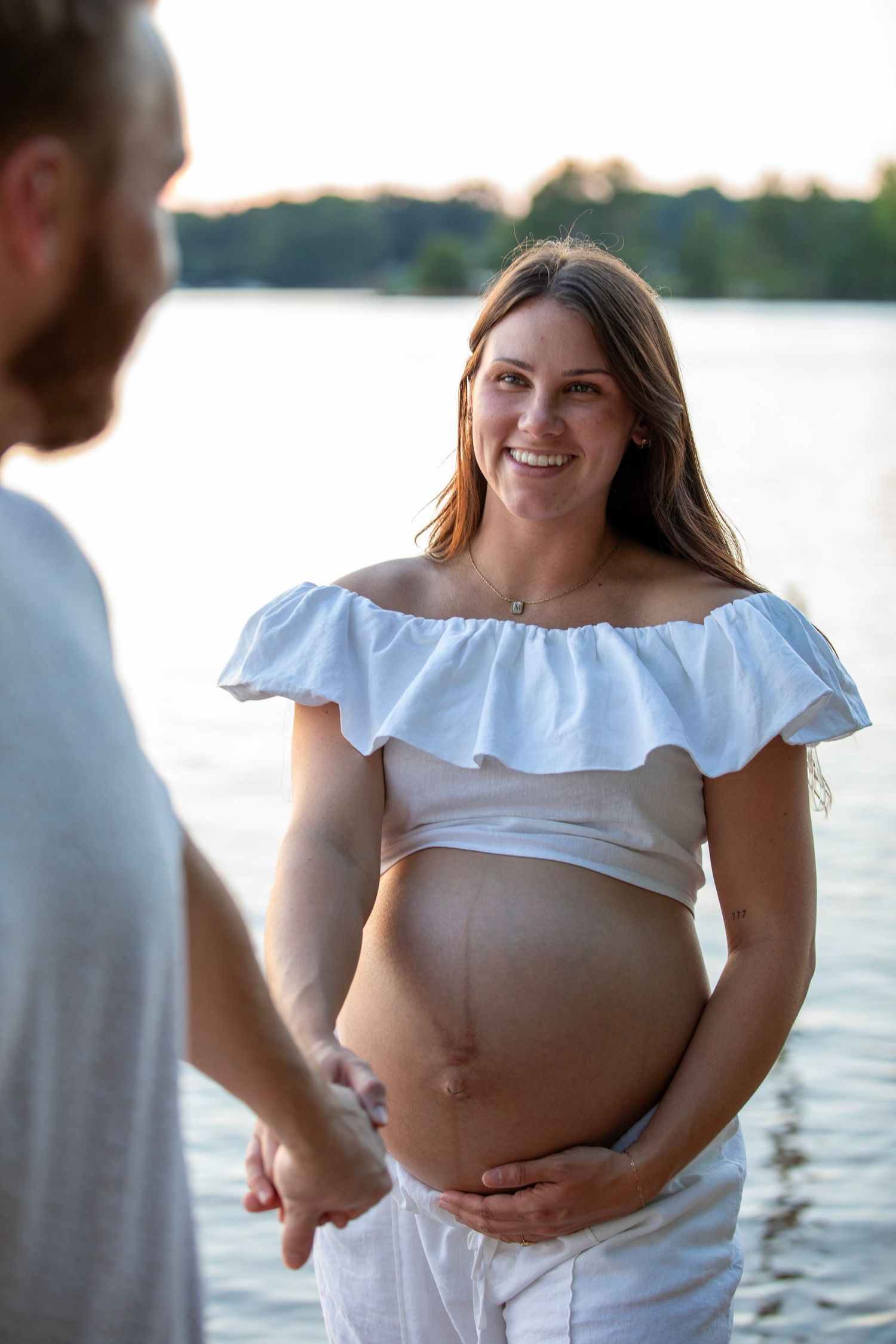 A maternity photoshoot showing a pregnant woman in a white off-shoulder top by a lake.