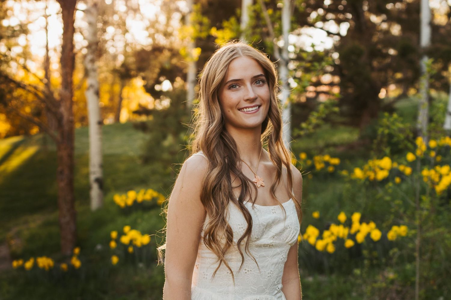 A young lady in white dress poses outdoors during golden hour among yellow wildflowers.