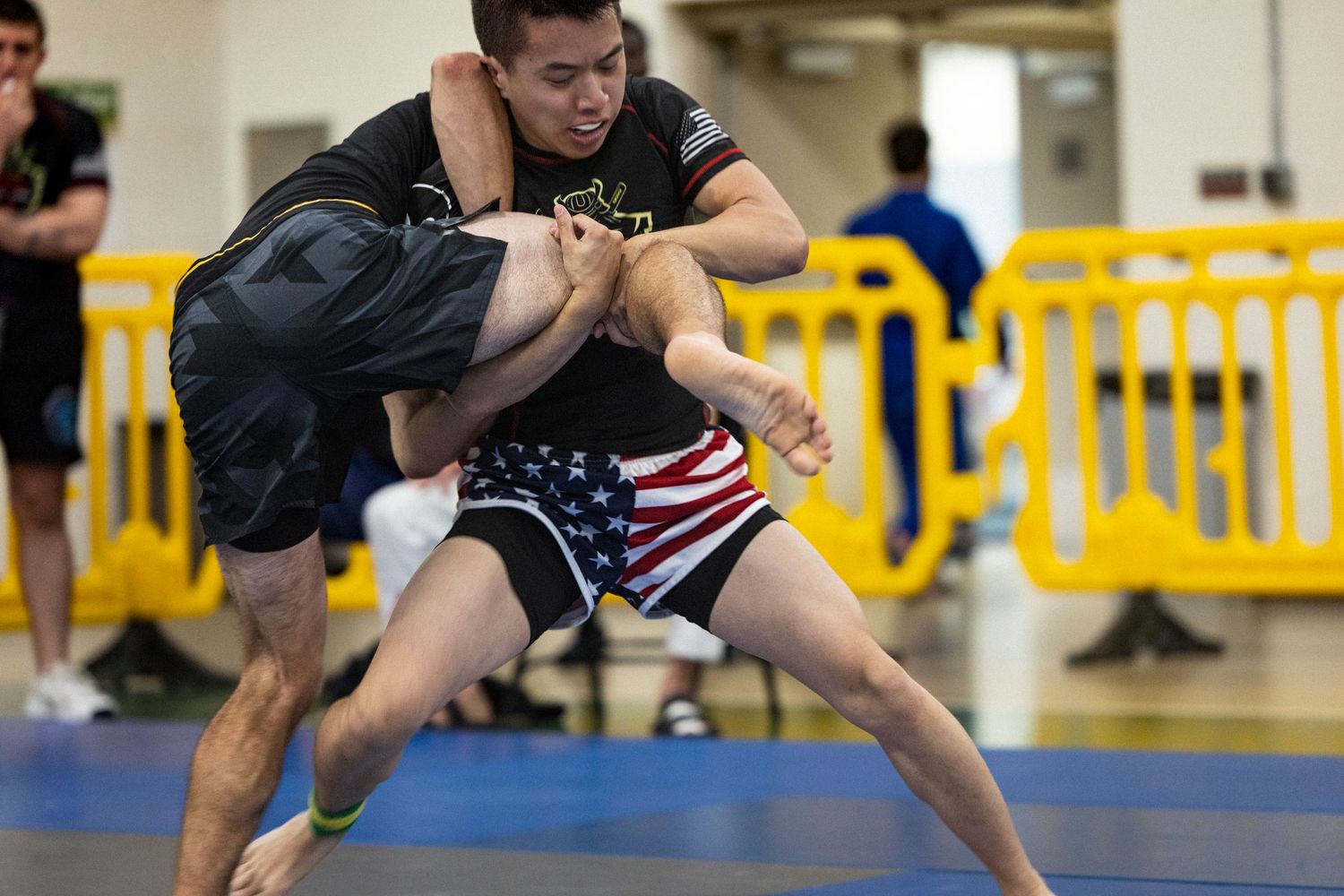 Two athletes grappling during a wrestling match with yellow barriers visible in the background.