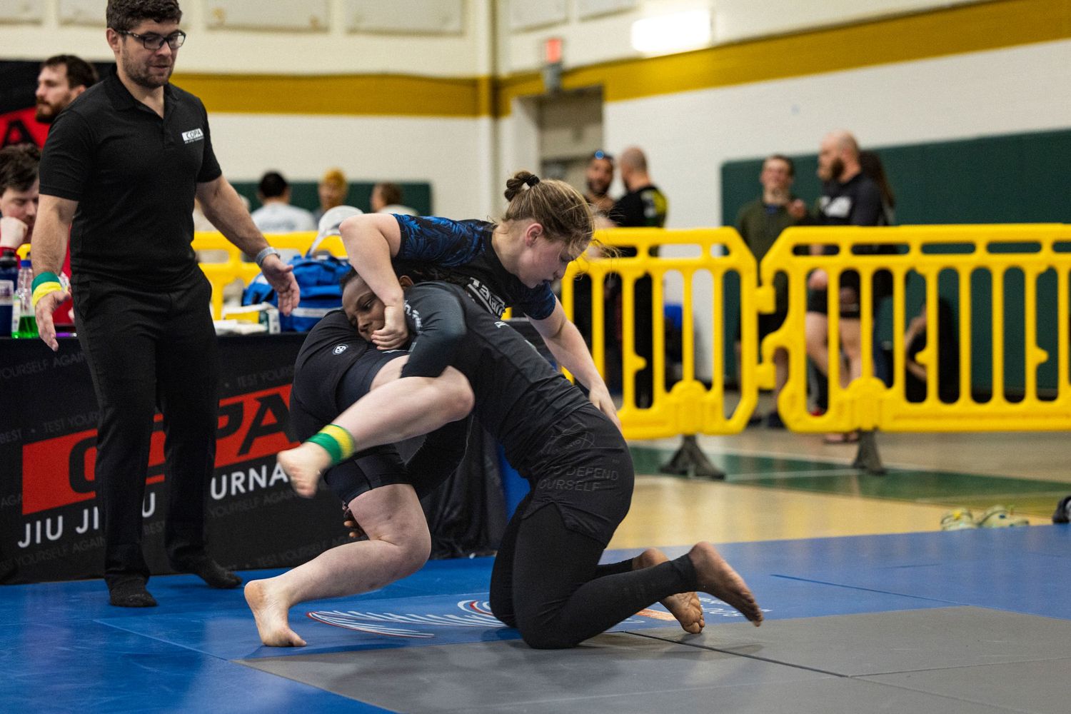 Two athletes compete on a blue mat during a jiu-jitsu tournament as officials observe from the sidelines.