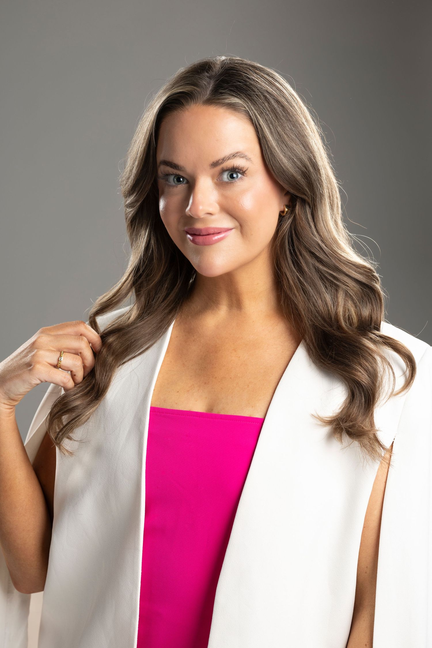 Studio portrait in a white blazer and pink top with flowing hair styled in loose waves.