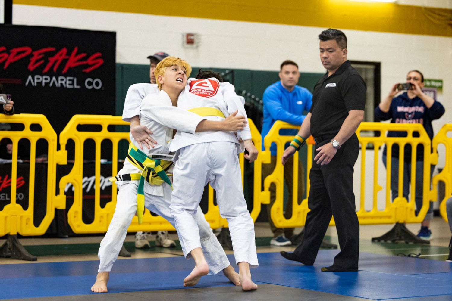Two martial artists in white gis grappling during competition as referee watches near yellow barriers on gymnasium floor.