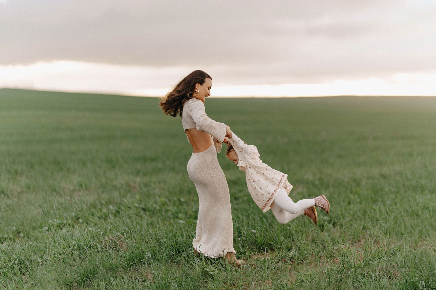 Two people in white dresses share a playful moment while spinning together in a green field under cloudy skies.