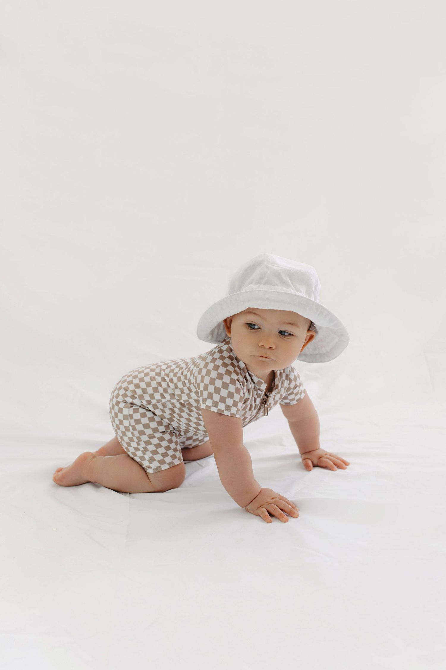 Baby wearing a white hat and gingham romper crawls on white studio background.