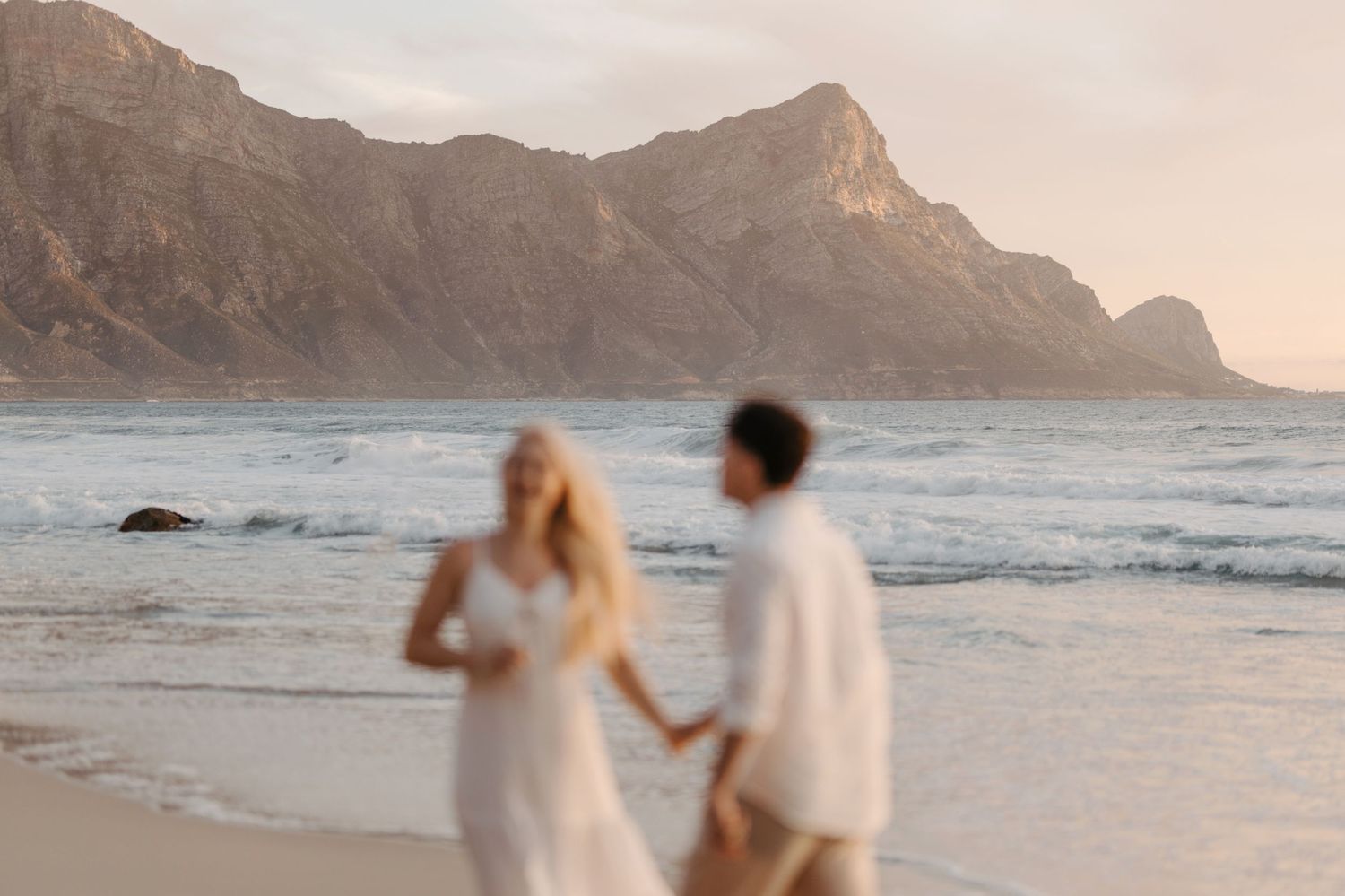 Couple walking on a scenic beach at sunset with dramatic mountain peaks in the background.