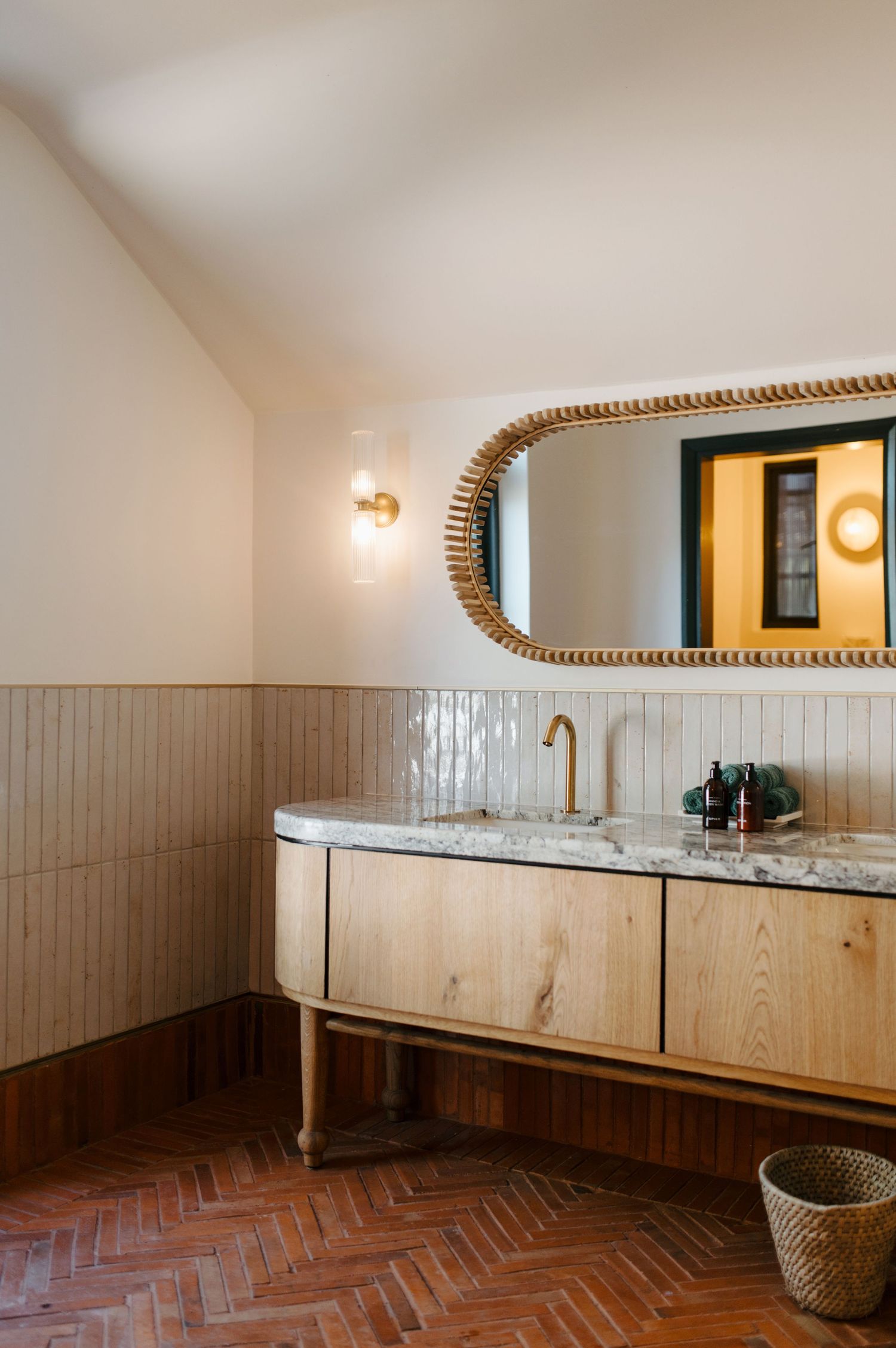 Modern bathroom interior with herringbone floor tiles, wooden vanity cabinet, marble countertop and oval gold-framed mirror.