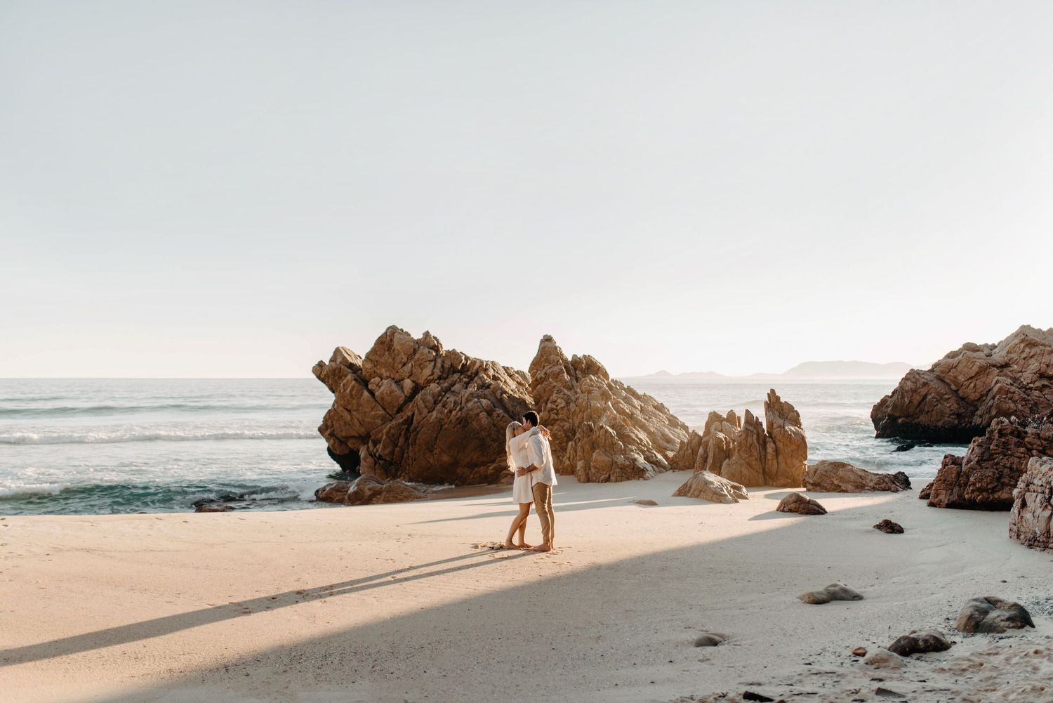 Scenic coastal landscape with rugged rock formations and sandy beach at sunset creating long shadows.
