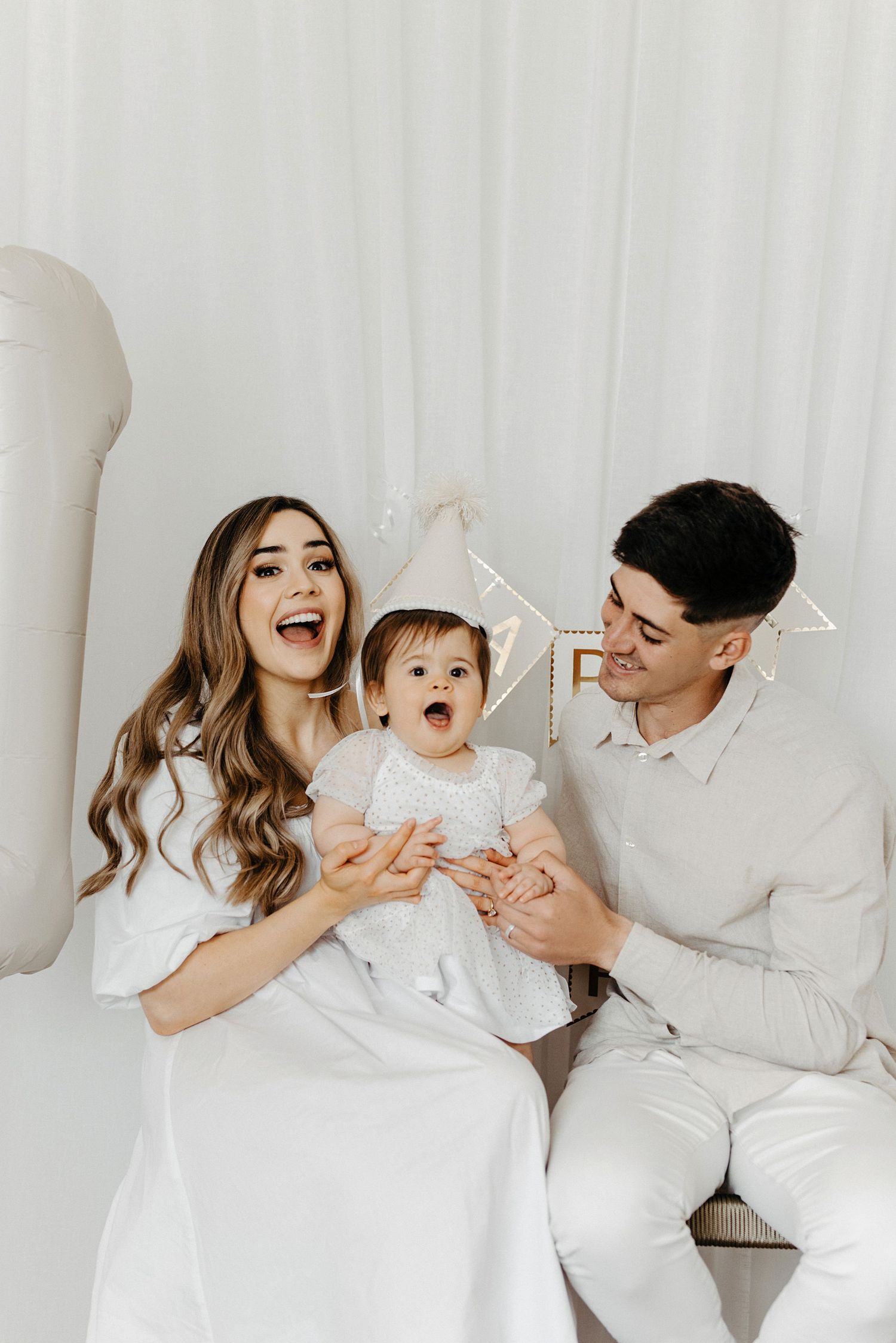 A family wearing white outfits sits together on a couch while smiling and laughing during a photo session.