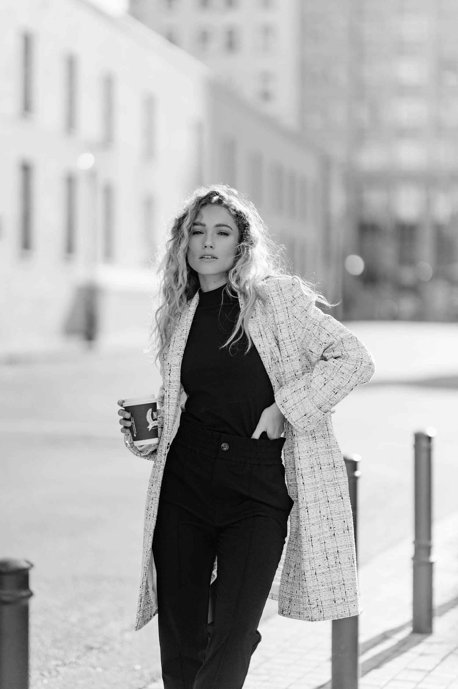 A fashionable woman in a tweed coat and black outfit poses confidently with a coffee cup on an urban street.