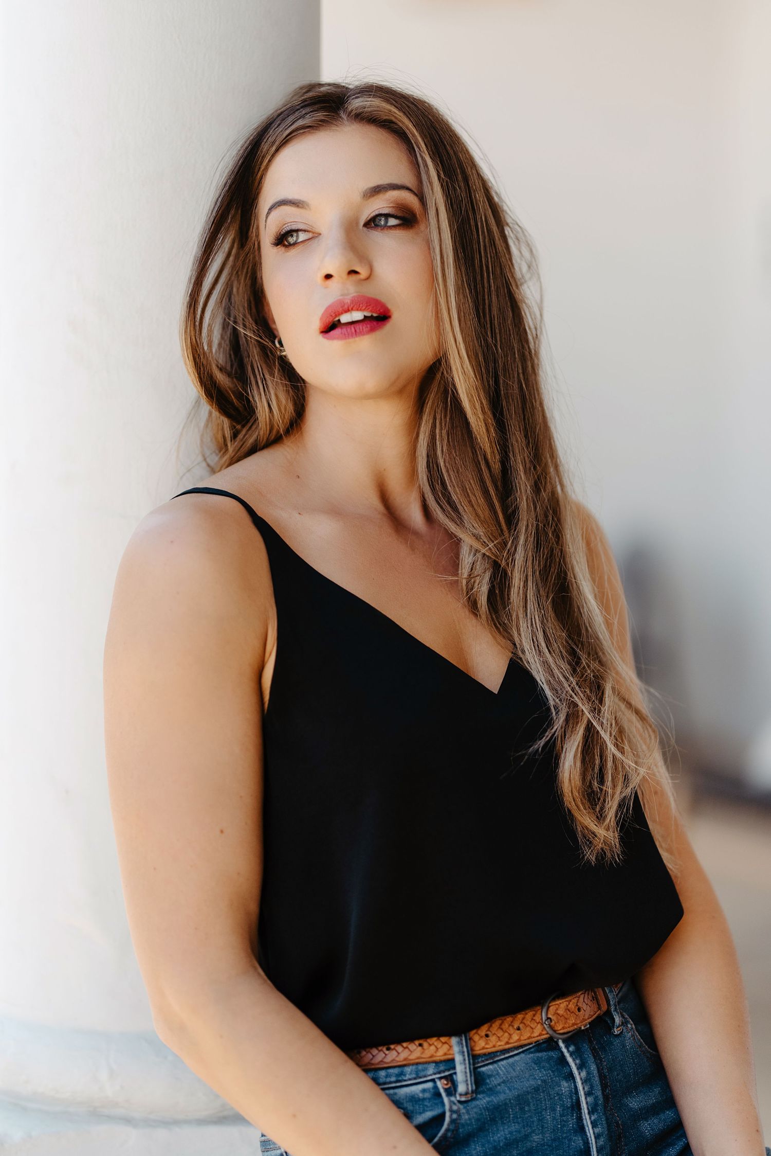 Woman with long brown hair wearing black tank top and jeans poses against white wall in casual fashion portrait.