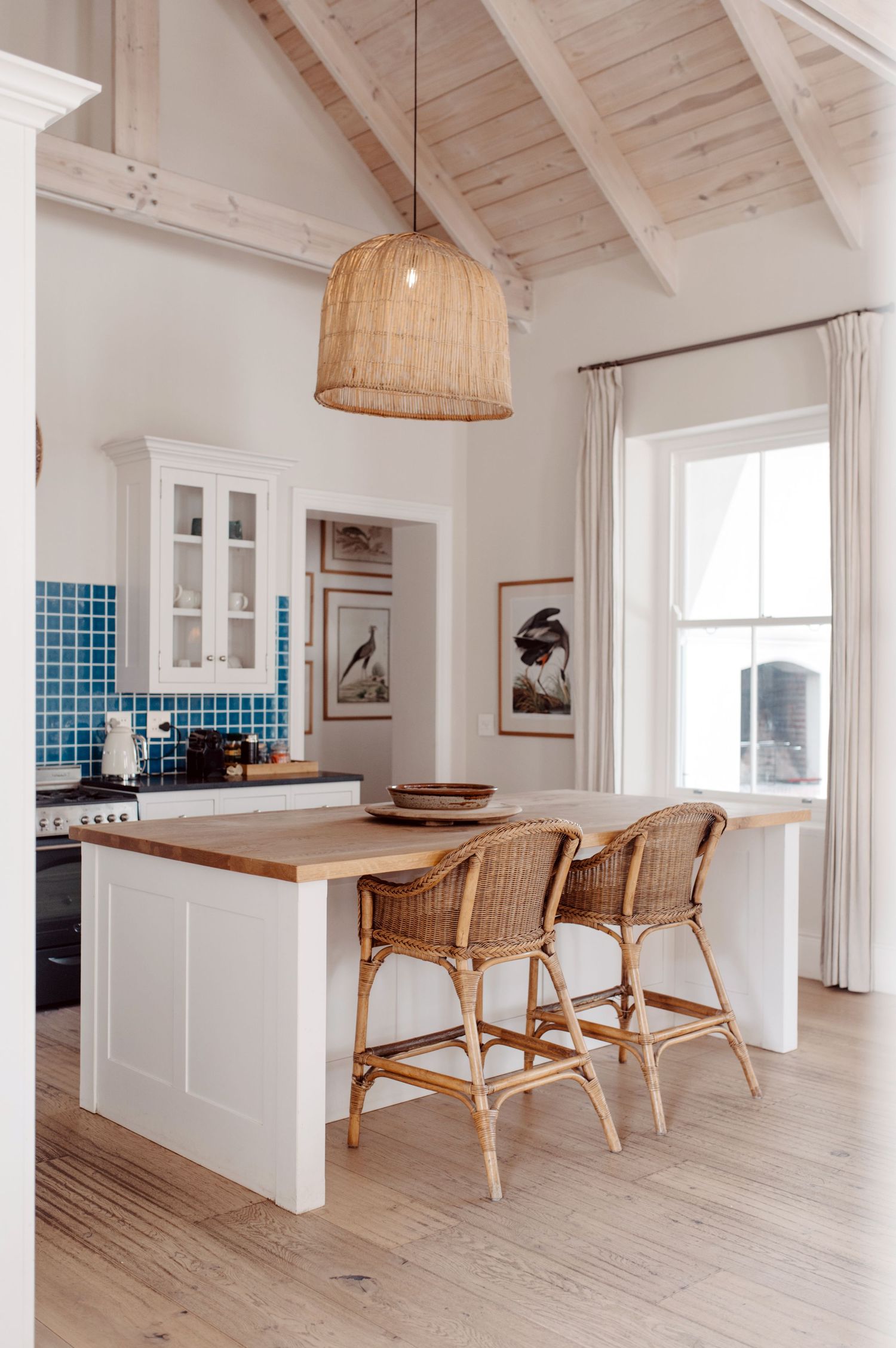 Modern white kitchen with blue tile backsplash, rattan chairs, and natural wood flooring in a bright coastal design.
