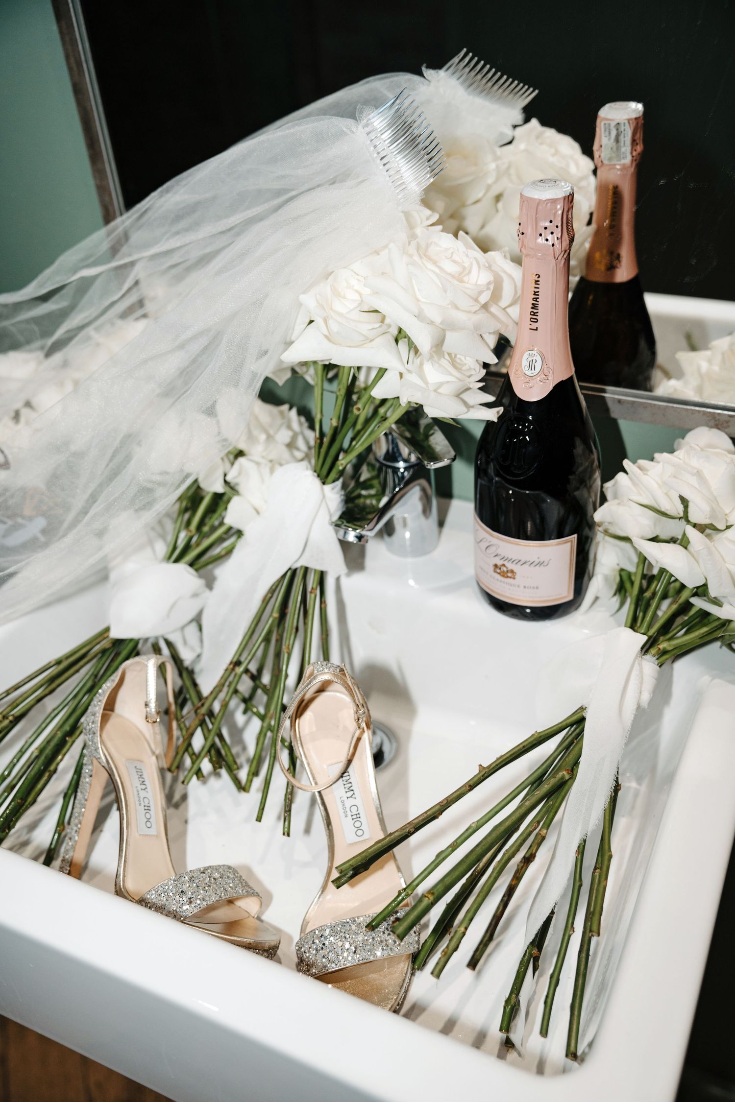 Wedding accessories including champagne, white heels, bouquet and veil arranged on white surface.