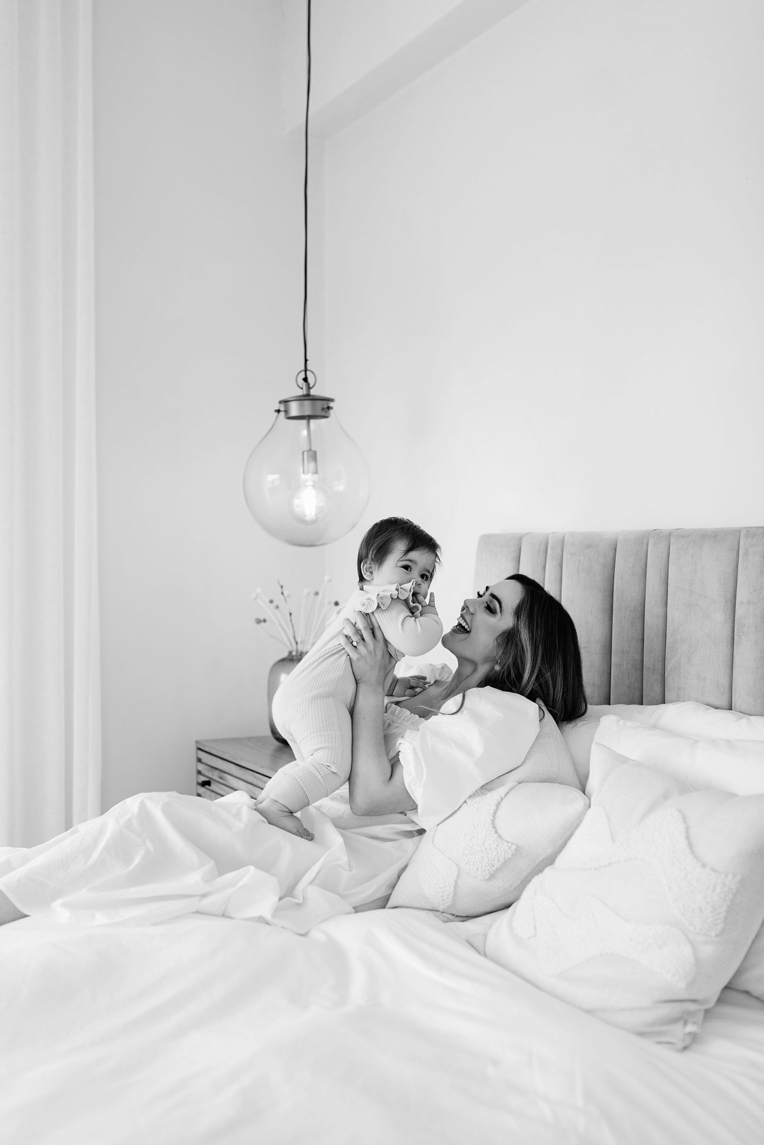 A tender moment captured in black and white as two people share affection on a white bed beneath a hanging pendant light.