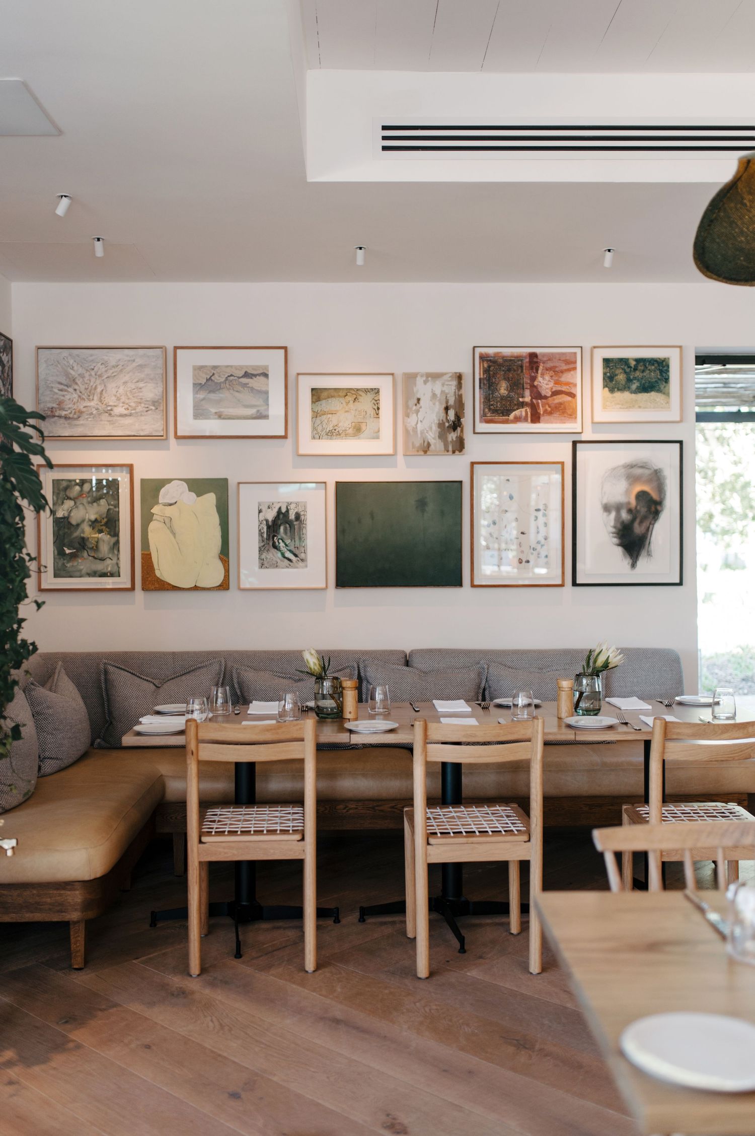 Modern restaurant interior with wooden tables, grey banquette seating, and framed artwork along white walls displaying a minimalist design.