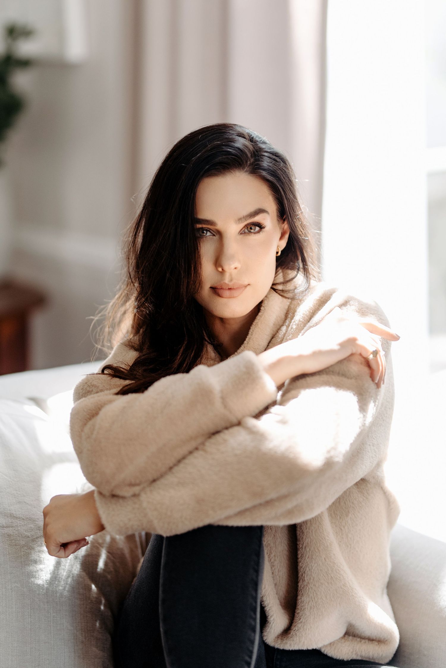 Portrait of a person in a beige sweater leaning forward and looking thoughtfully into the distance against a white wall with sunlight.