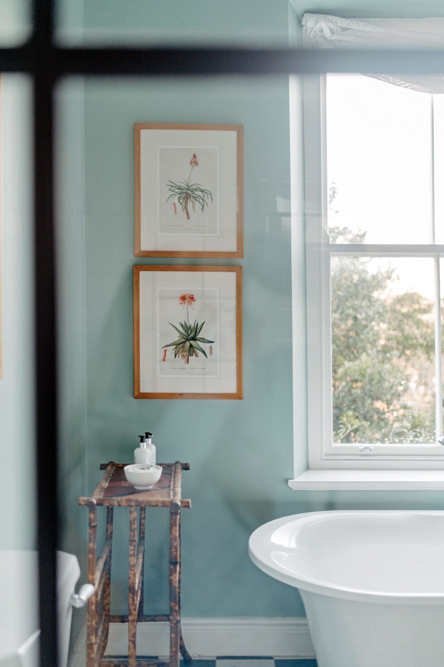 Modern bathroom with soft blue-green walls featuring a freestanding tub, wooden stool, and framed botanical prints.