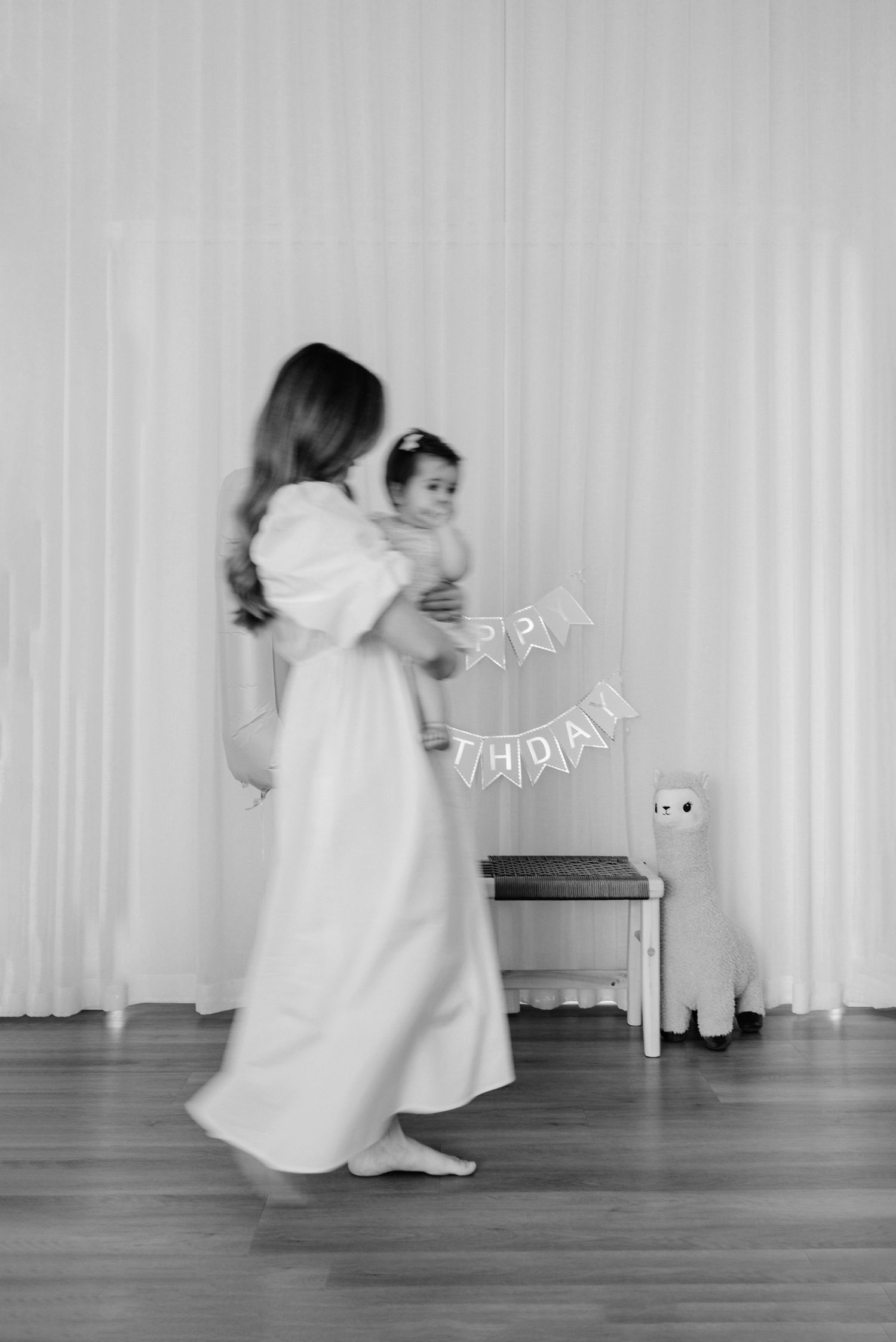 A person in a long white gown dances with a baby in a nursery setting with banner decorations.