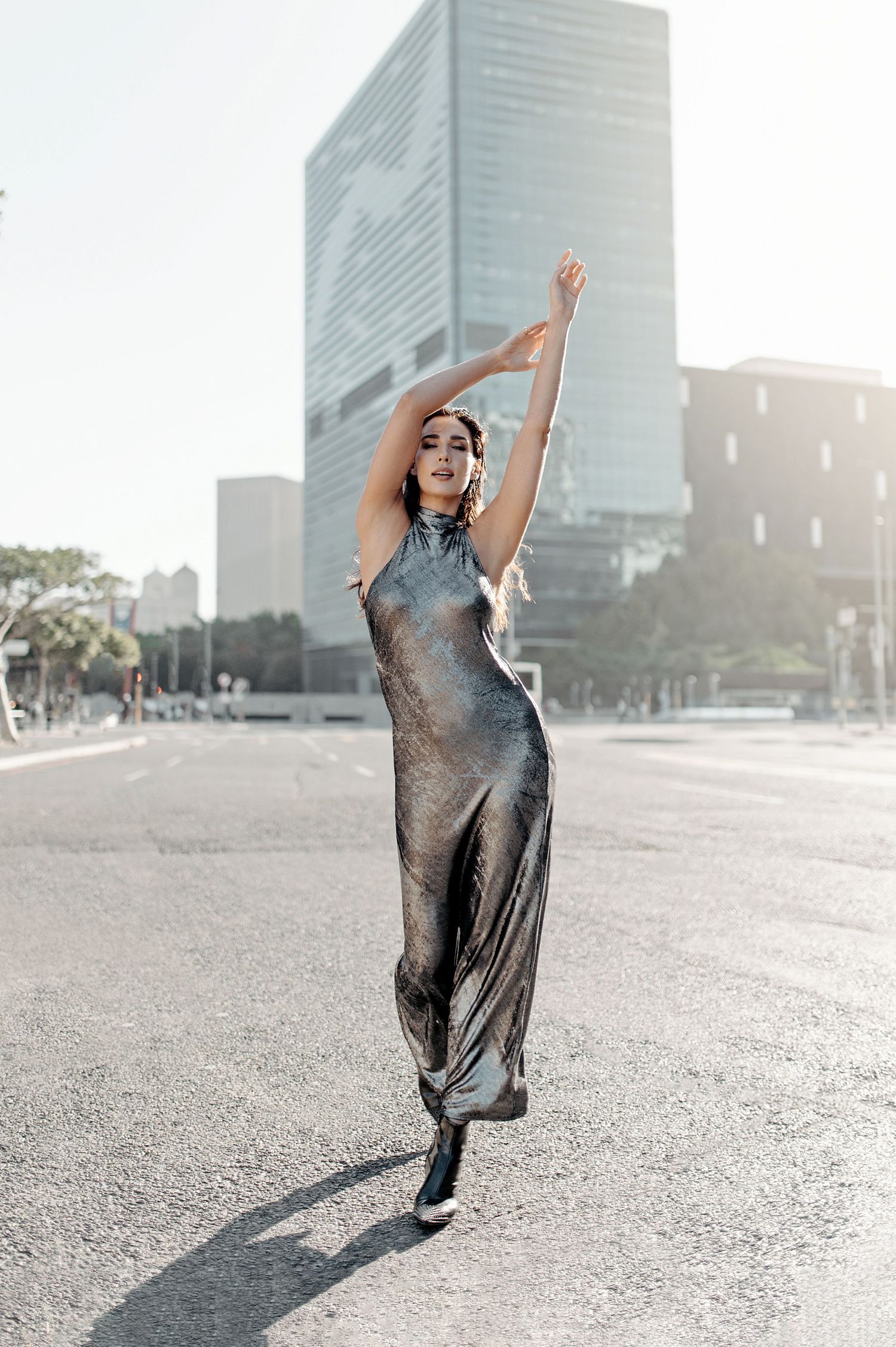 A graceful dancer in a metallic dress poses elegantly on a city street with modern buildings in the background.