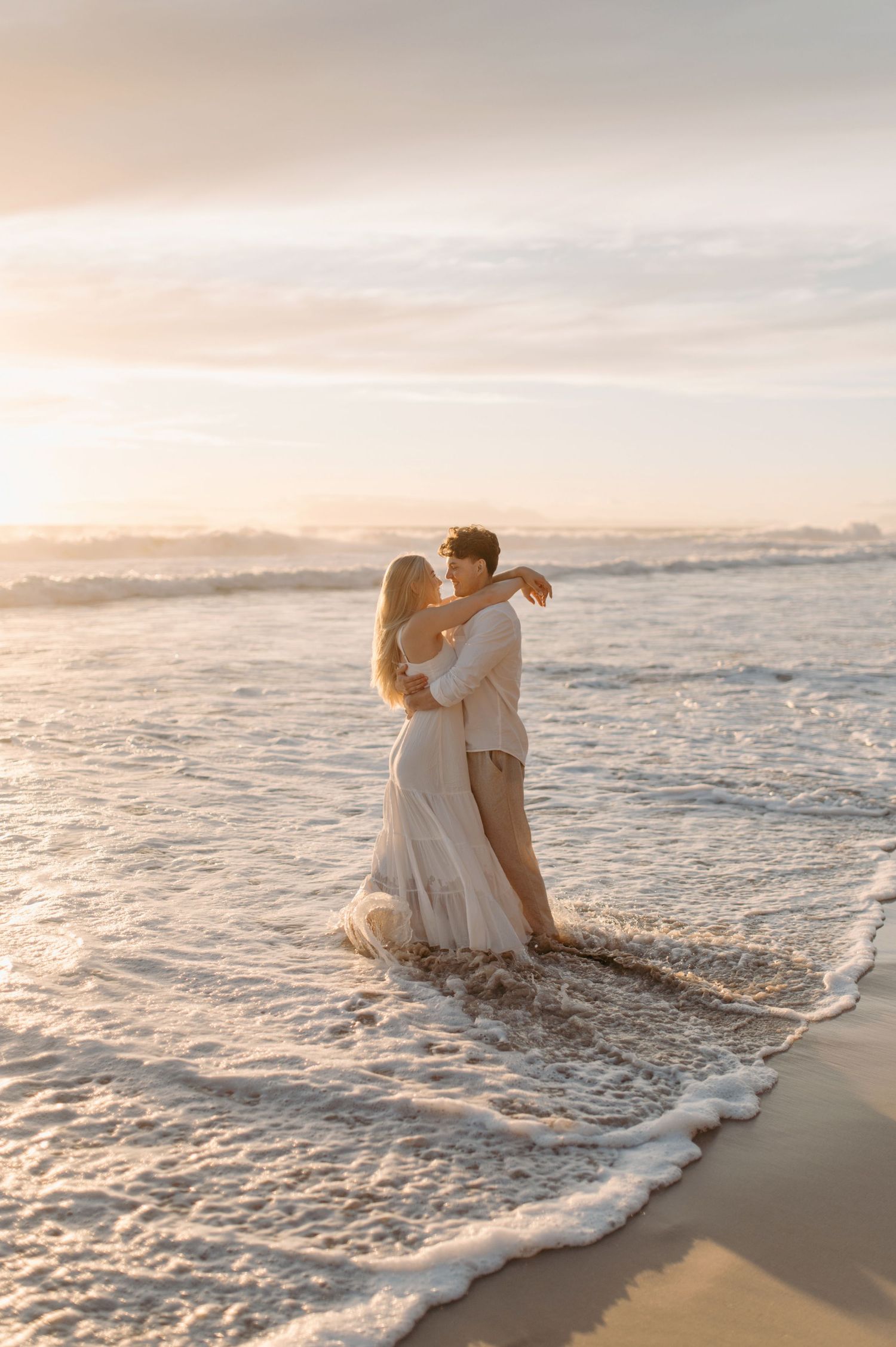 A romantic beach scene at sunset with a couple embracing in formal attire as waves lap at their feet.