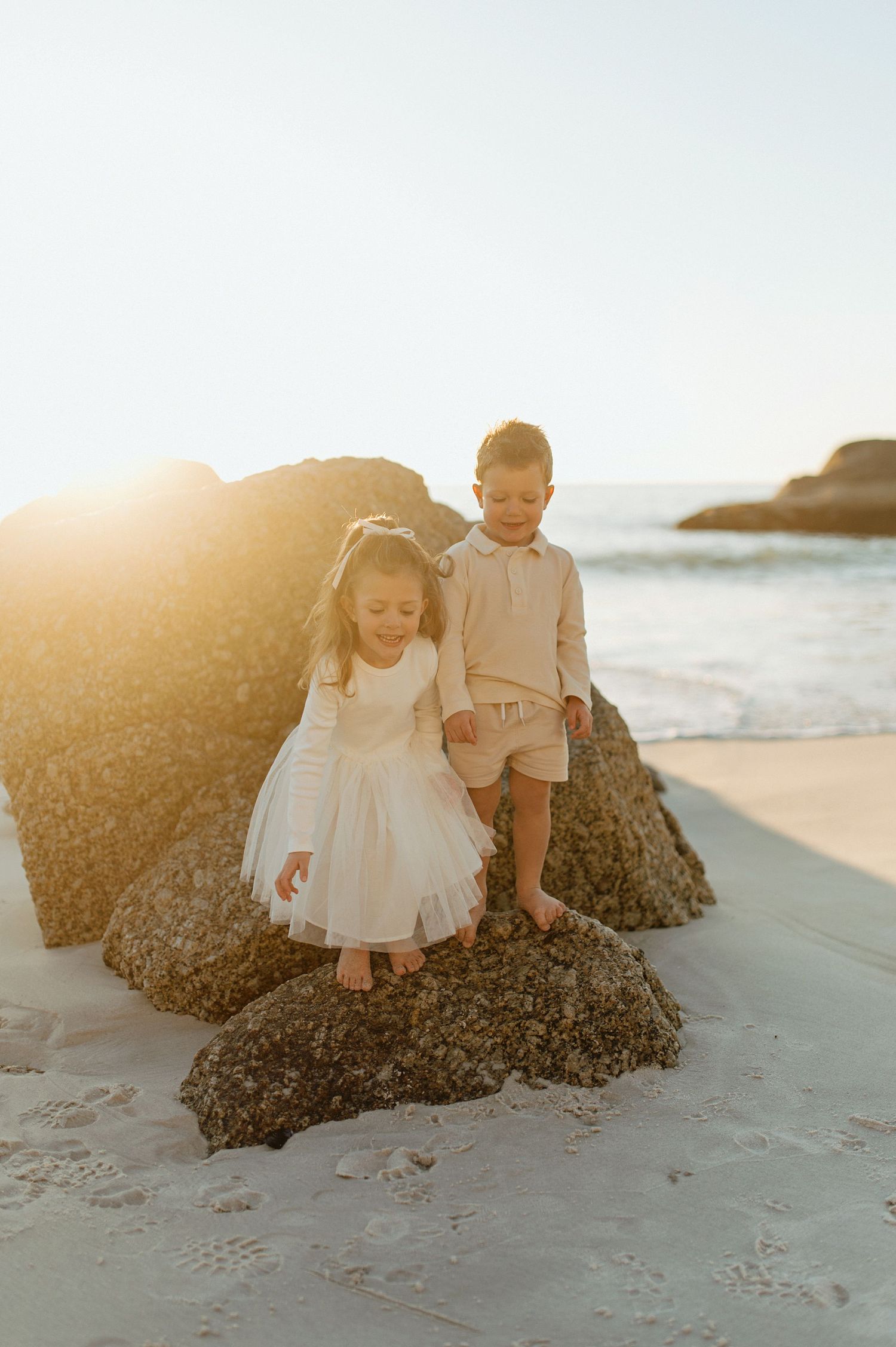 Two children stand atop rocky beach formations during golden hour sunset as waves crash along the shoreline.