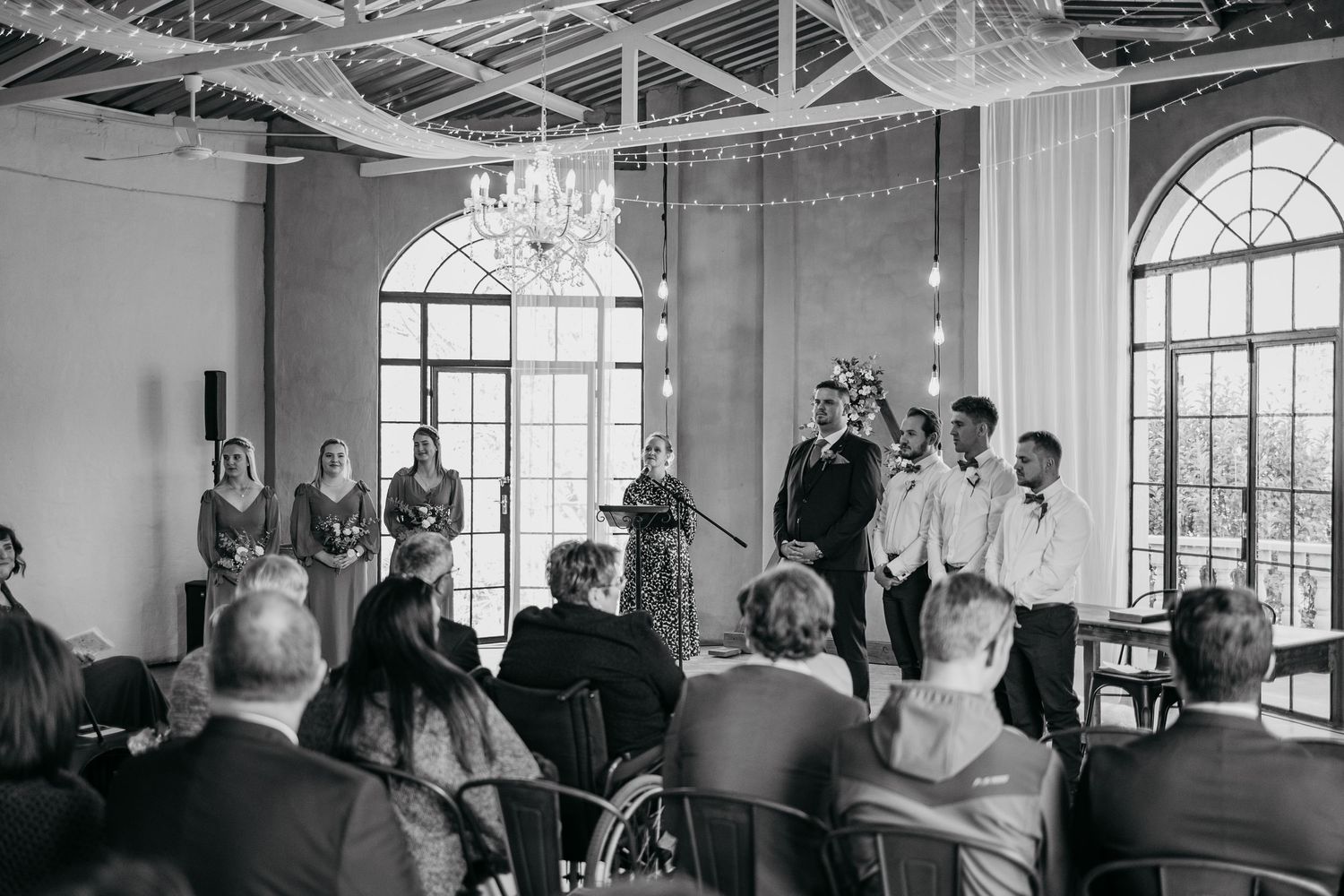 A black and white photograph of a wedding ceremony in an elegant room with arched windows and a chandelier.